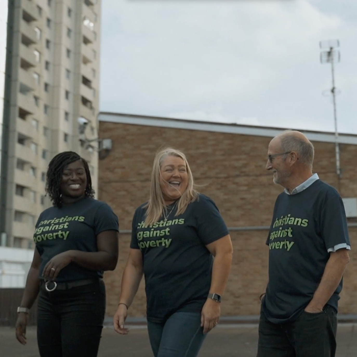 Three people wearing black t-shirts with green text that reads 'Christians Against Poverty,' walking together and smiling in an outdoor urban setting with a tall apartment building and a brick wall in the background.