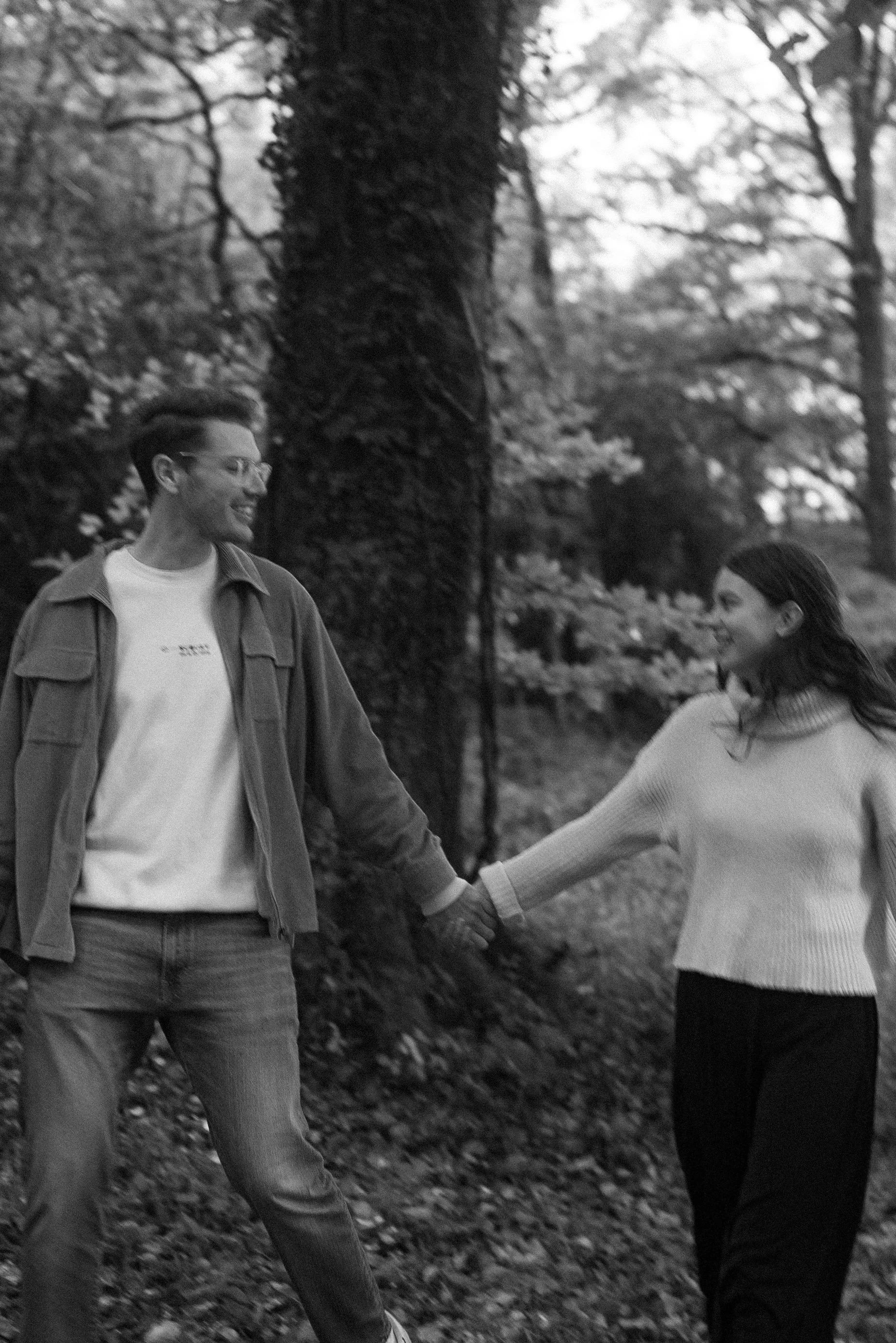 Two women holding hands and smiling at each other while walking through a wooded area with fallen leaves in the background.