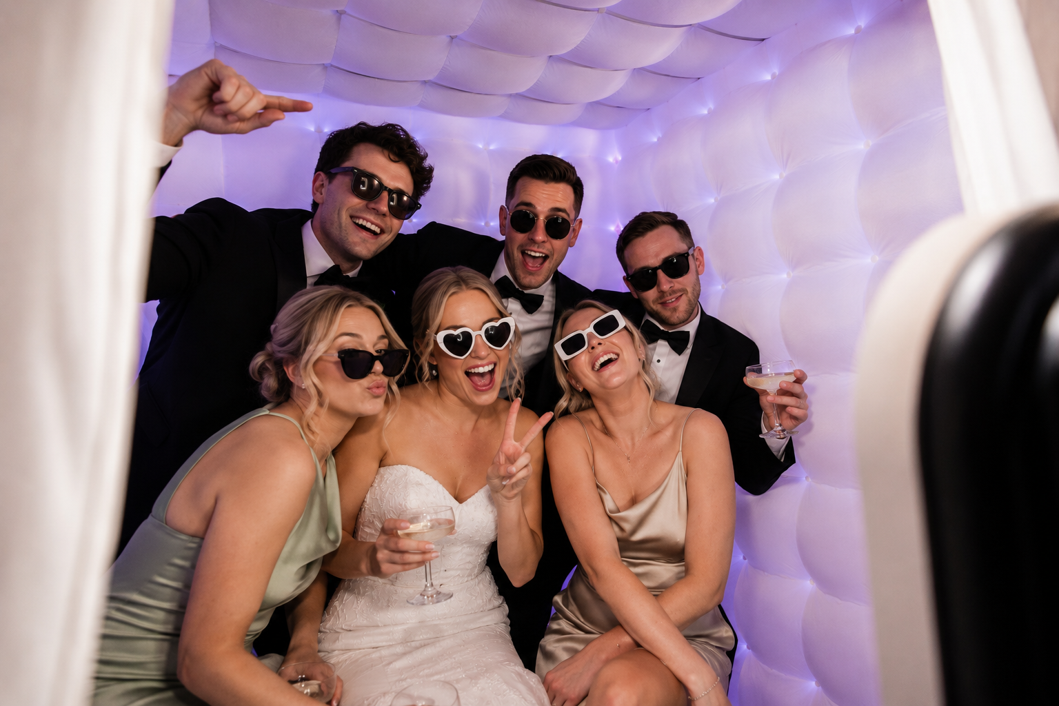Group of friends dressed in formal attire and sunglasses taking a selfie in a photo booth at a celebration, with some holding drinks.