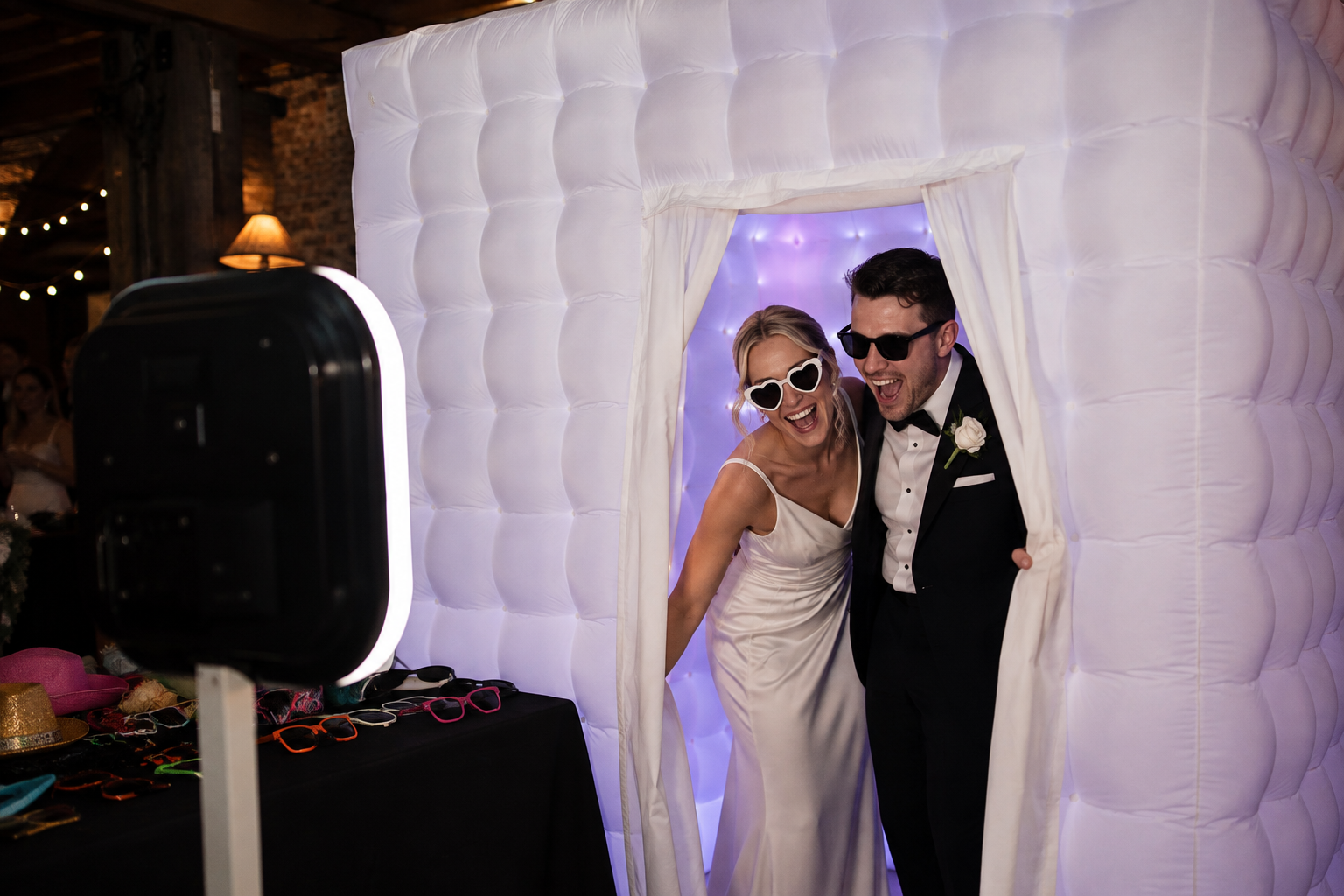 A bride and groom wearing sunglasses laugh and smile as they pose in a photo booth at their wedding reception. The booth has a white, cushioned, tufted outer wall and a purple-lit interior.