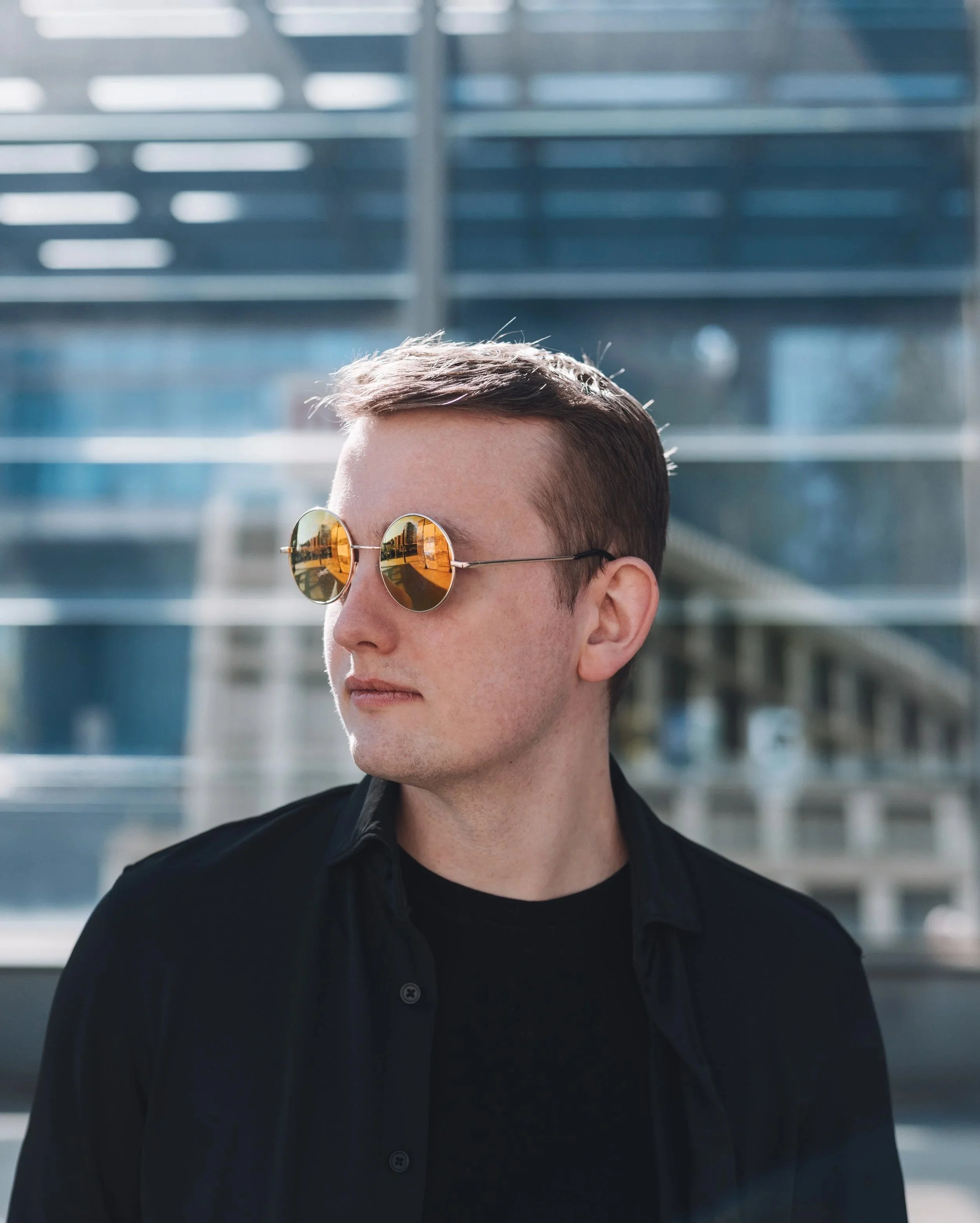 Close-up of a young man with short brown hair wearing sunglasses with orange-tinted lenses, a black button-up shirt, standing outdoors with a blurred metal structure in the background.