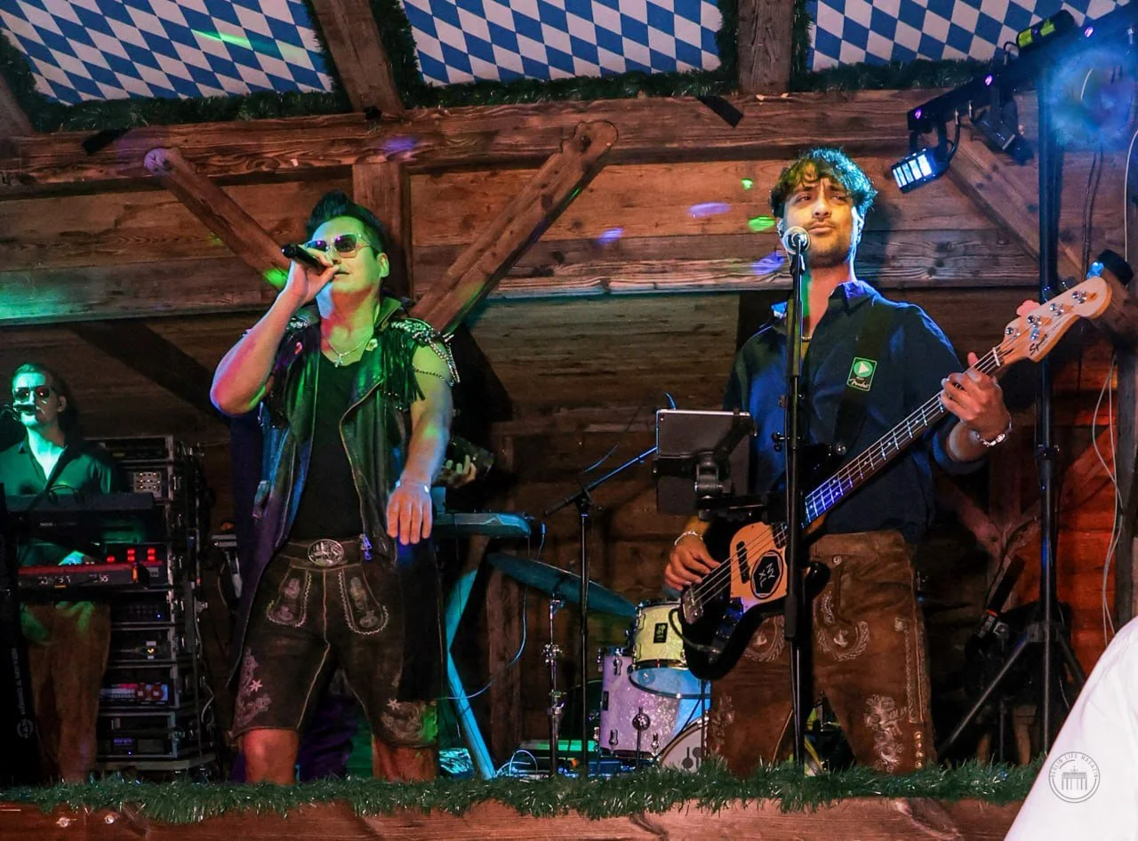 A band performing on stage with wooden background and German flag decorations. The lead singer, wearing sunglasses and a leather vest, is singing into a microphone. A bassist in a black shirt and traditional leather pants is playing guitar, while ano
