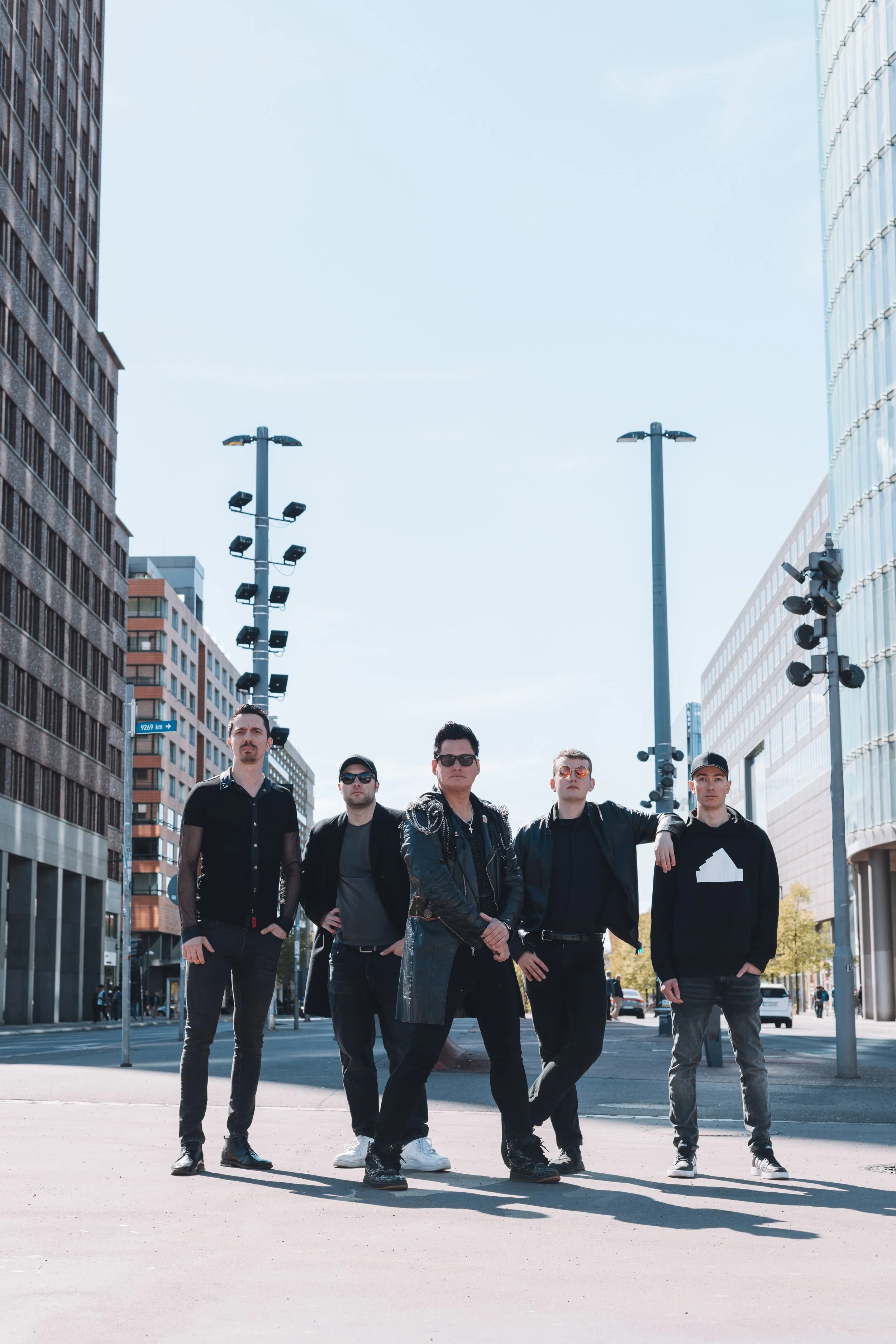 A group of six young men standing in the middle of a city street, posing confidently for a photo. The background features tall modern buildings, streetlights, and a clear sky.