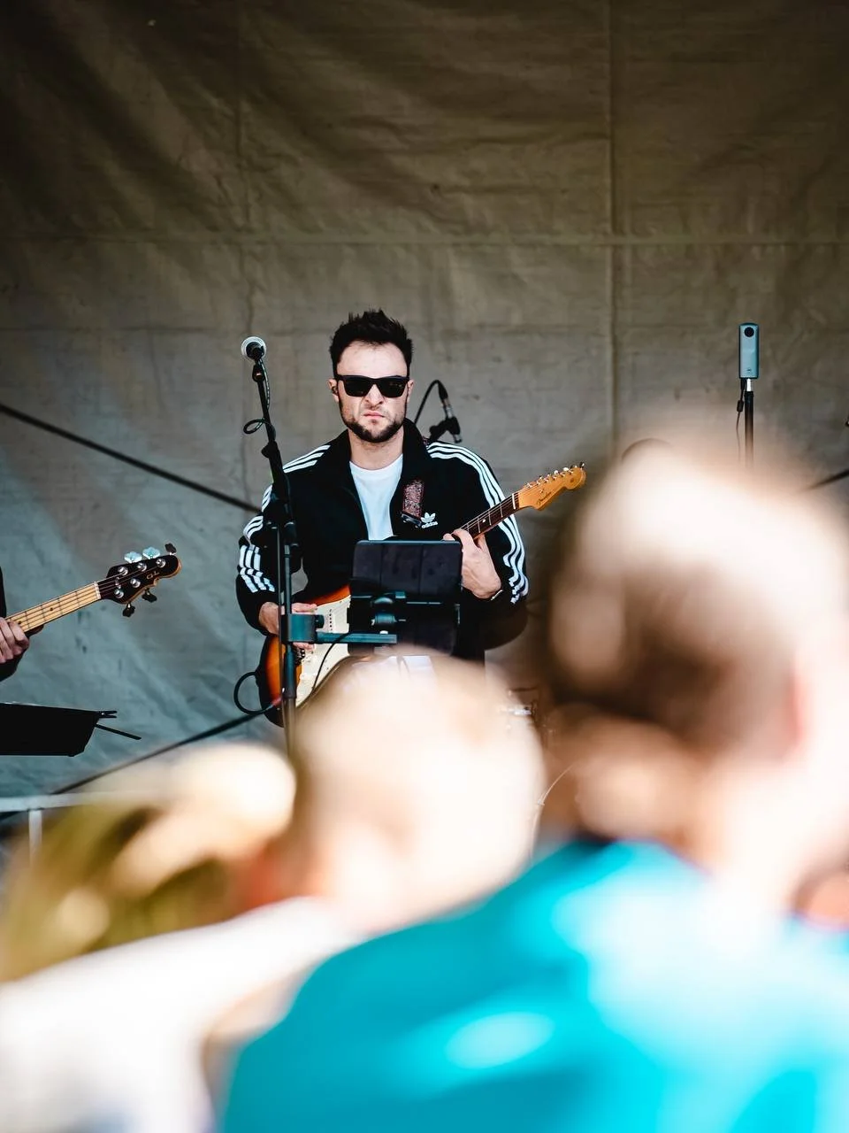 Musician playing electric guitar in front of an audience, wearing sunglasses and a black Adidas jacket with white stripes, with other band members and audio equipment visible in the background.