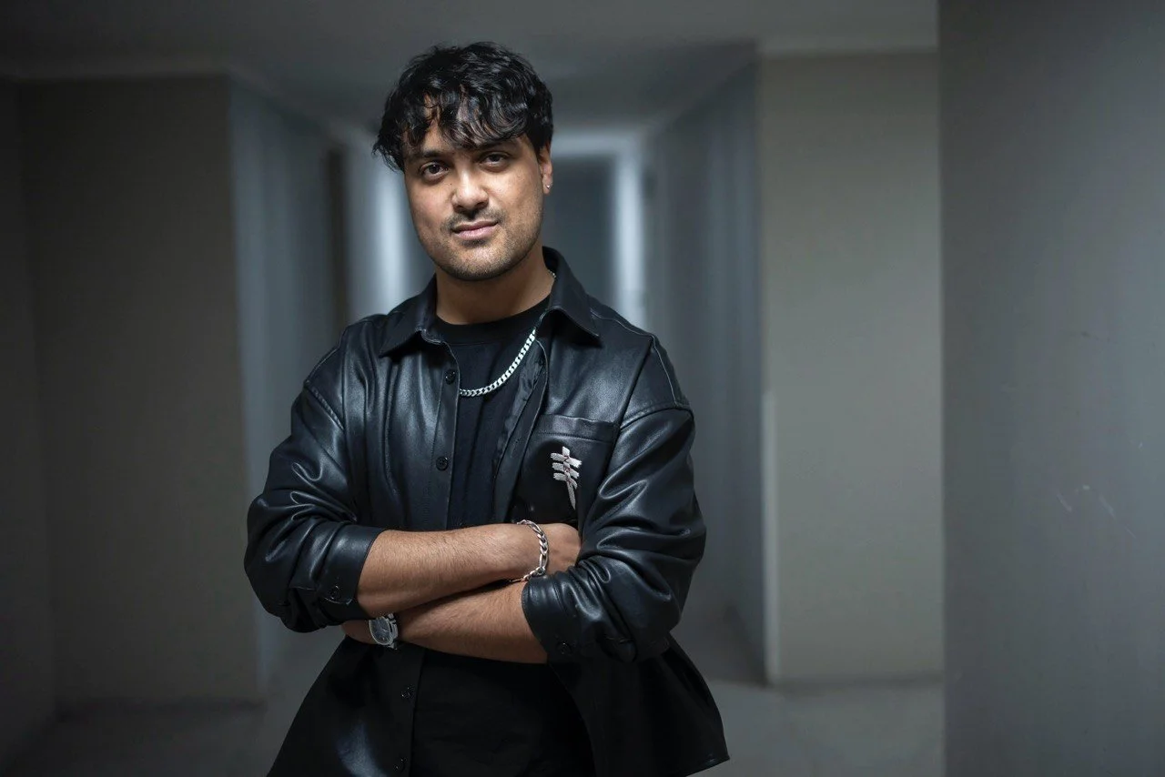 Young man with dark curly hair, wearing a black leather jacket, a black shirt, silver chain necklace, and watch, standing with arms crossed in a dimly lit corridor.