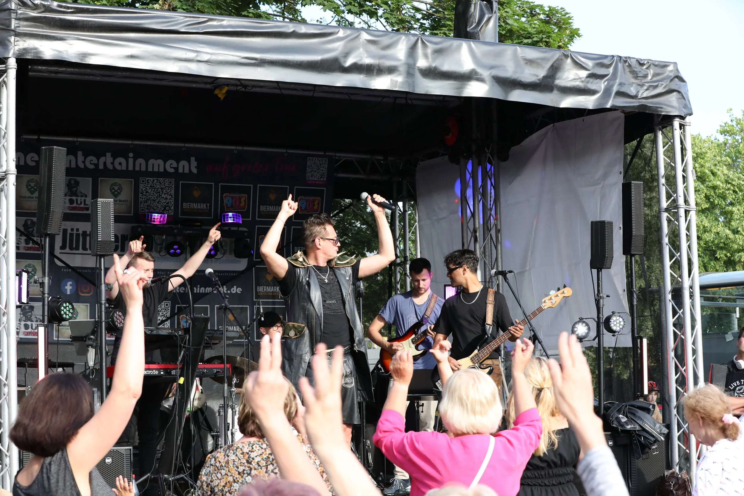 A band performing on an outdoor stage with several people in the audience raising their hands and cheering. The band members are playing instruments and the stage is equipped with speakers, lights, and banners.
