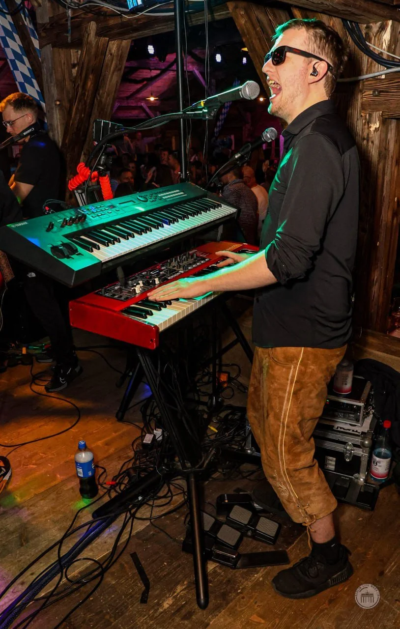 A male musician singing and playing a keyboard at a live music venue. He is wearing sunglasses, a black shirt, and brown pants. There is a crowd of people in the background enjoying the performance.