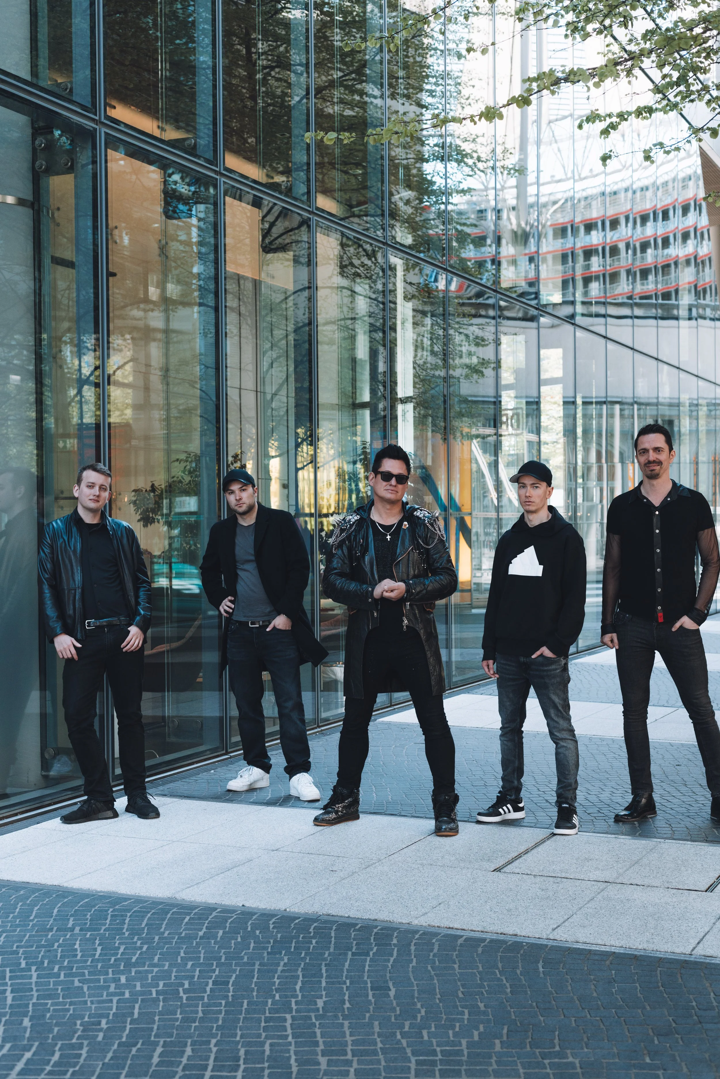 Five young men standing outdoors in front of a glass building with reflections of trees and a Ferris wheel in the background. They are dressed in casual and stylish clothing, with some wearing sunglasses and hats.