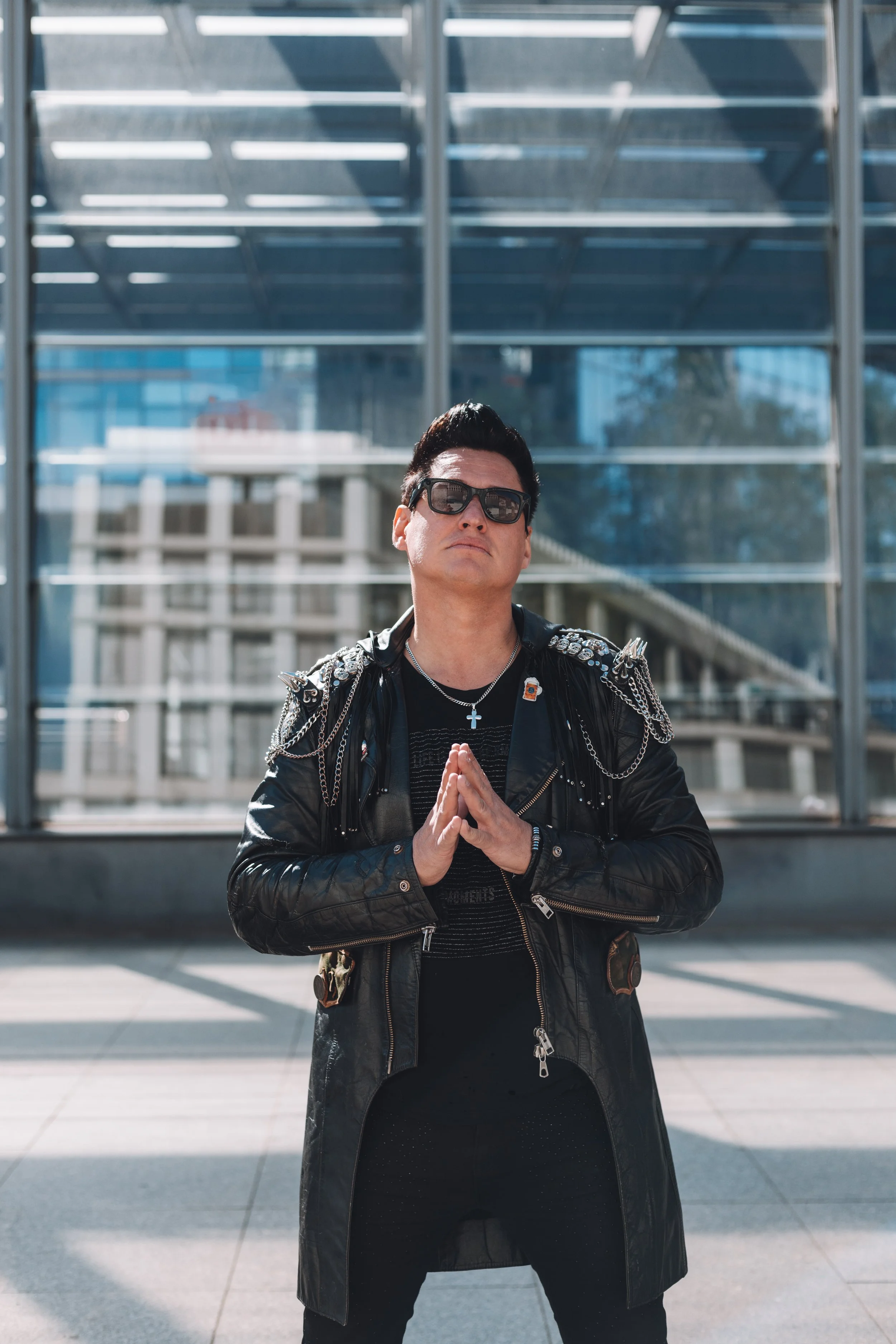 A man with black hair, sunglasses, and a leather jacket with metal spikes standing outdoors with hands in a prayer position. Modern glass building in the background.