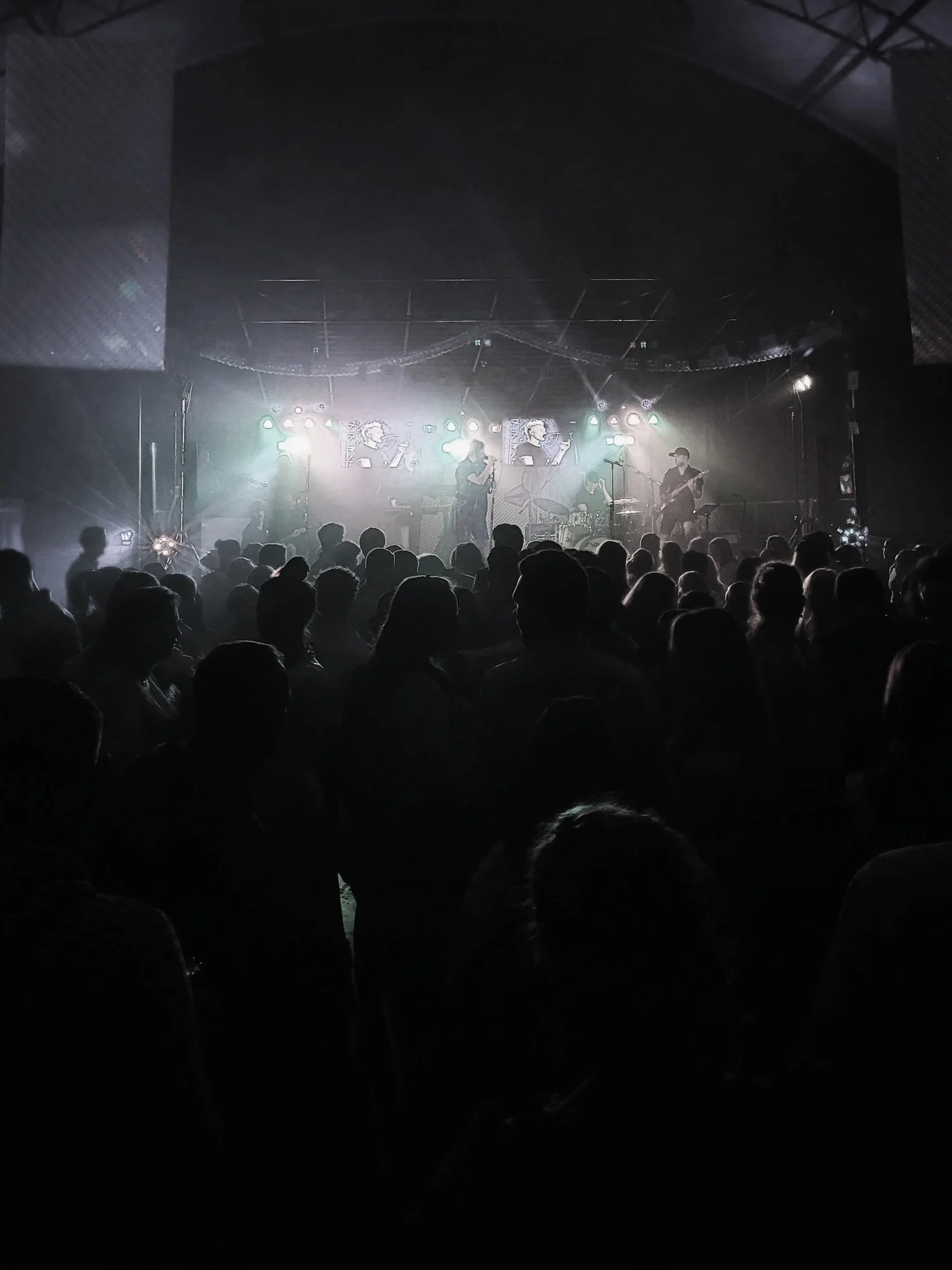 A live music concert with a band playing on stage, audience in silhouette, stage lights, and screens displaying musicians.