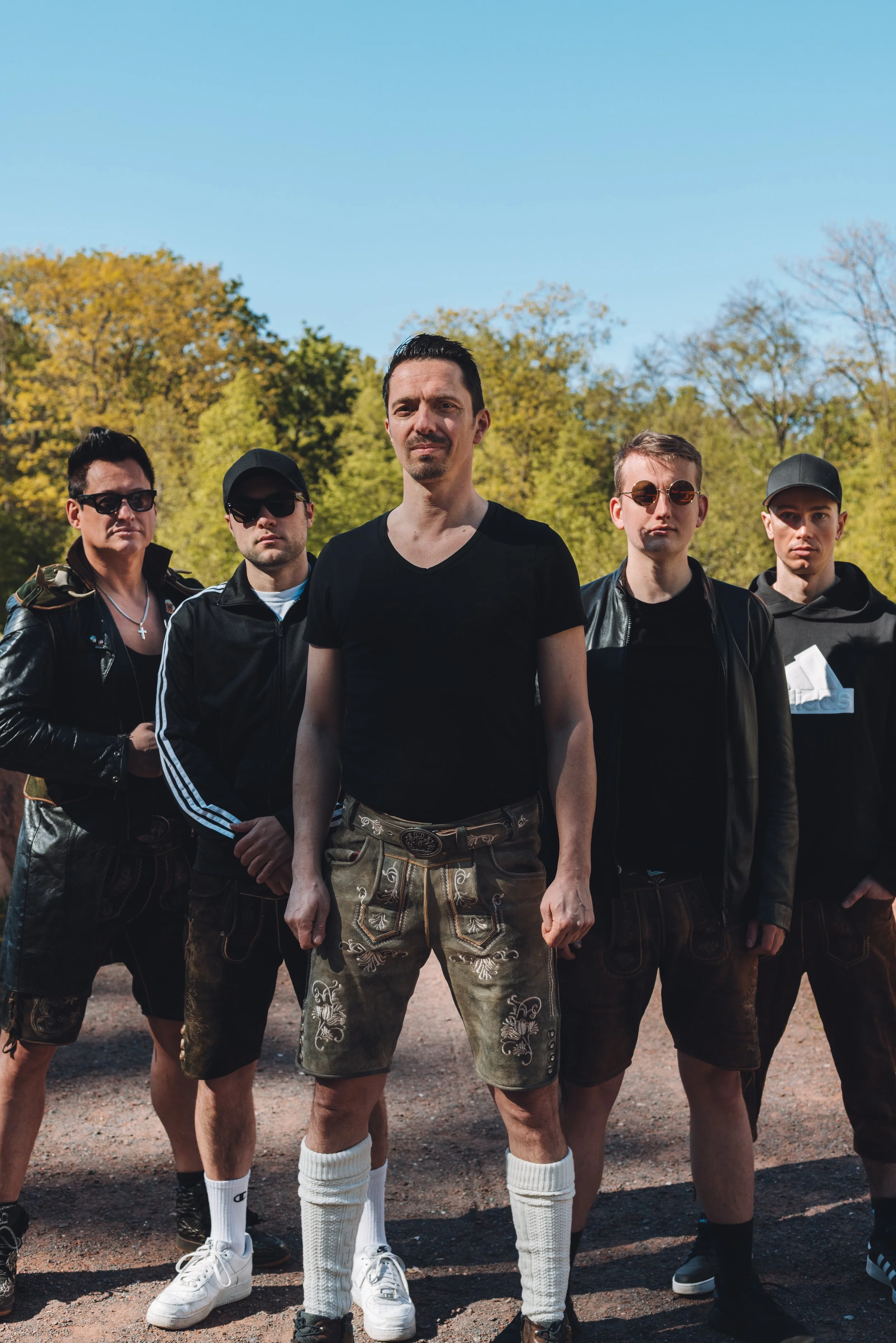 A group of five men standing outdoors on a sunny day with trees in the background. They are dressed in casual and sporty clothing, some with sunglasses and hats.