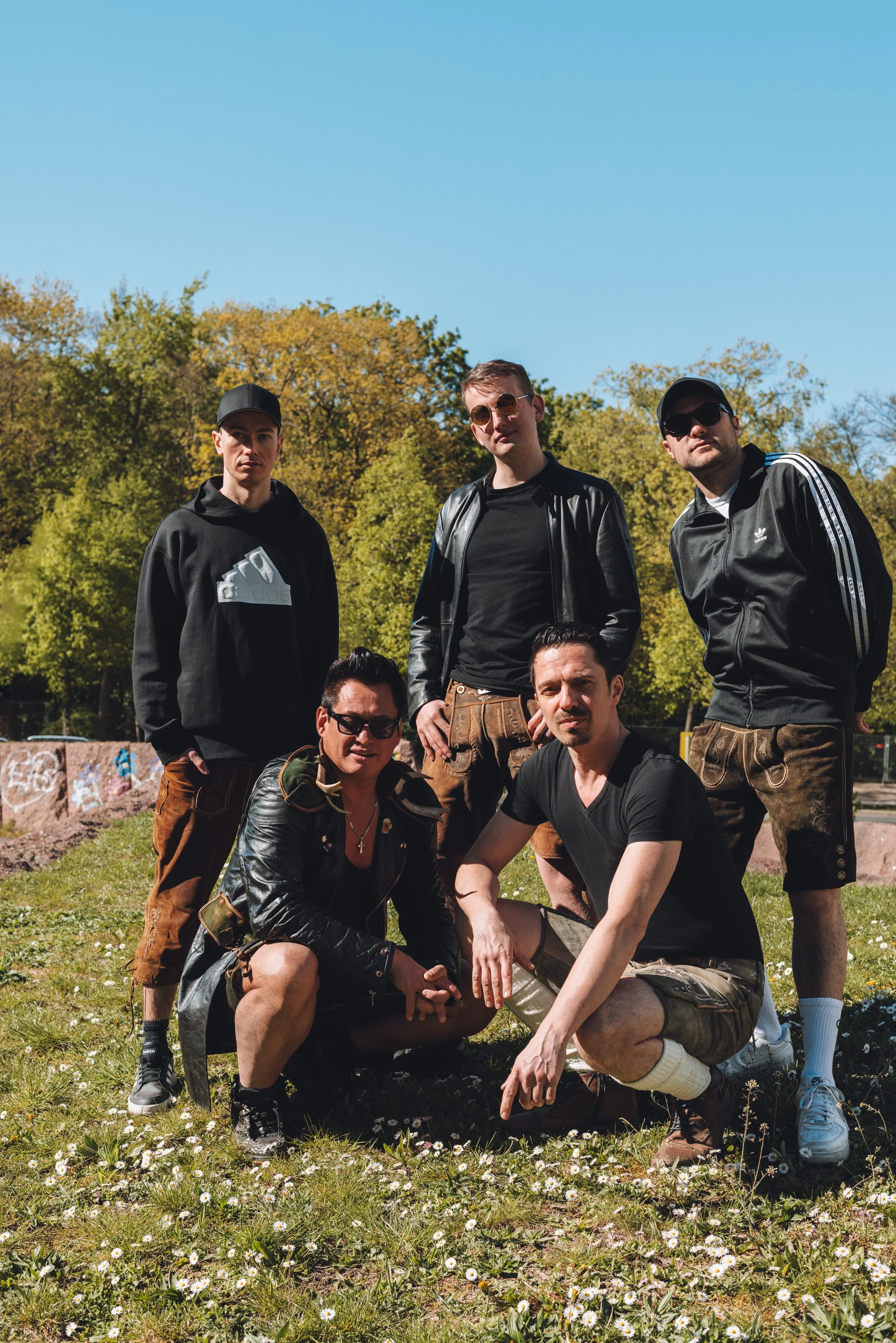 Group of five young men outdoors on a sunny day, standing and kneeling among small white flowers and green grass, with trees in the background.