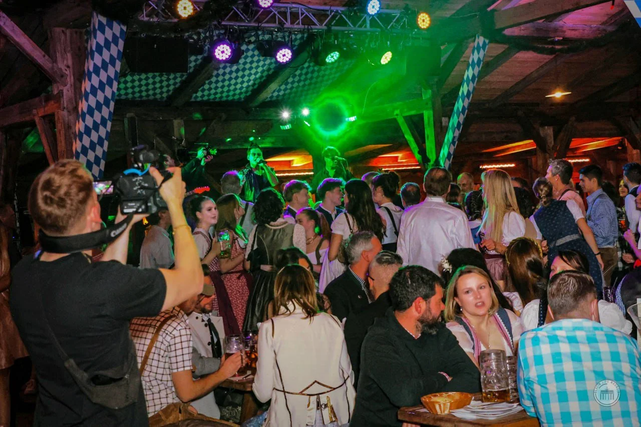 People gathered in a wooden hall for a celebration or concert, with some wearing traditional German clothing, and a band performing on stage with green lighting.