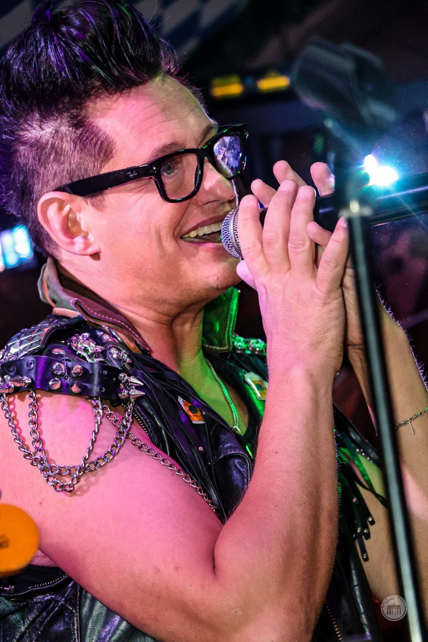 Person with short hairstyle, black glasses, and a studded leather jacket singing into a microphone during a performance.