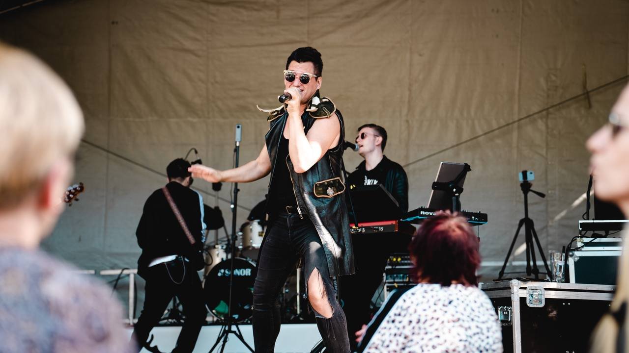 A male singer in sunglasses, a sleeveless leather jacket, and torn jeans performing on stage with a band, audience members watching.