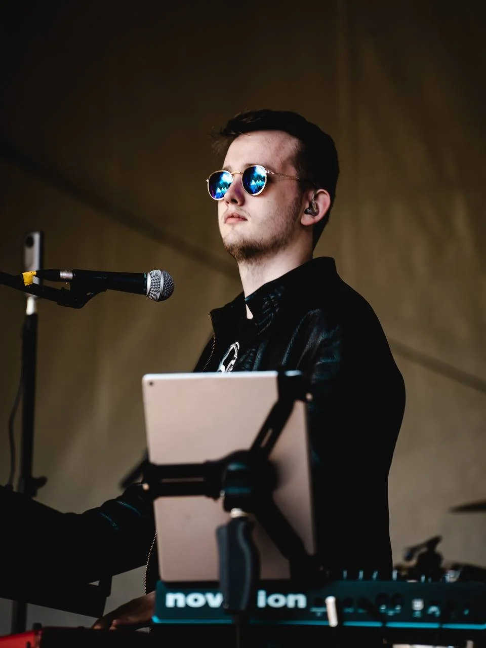 A young man with dark hair and sunglasses performing at a music event, standing behind a microphone and equipment.