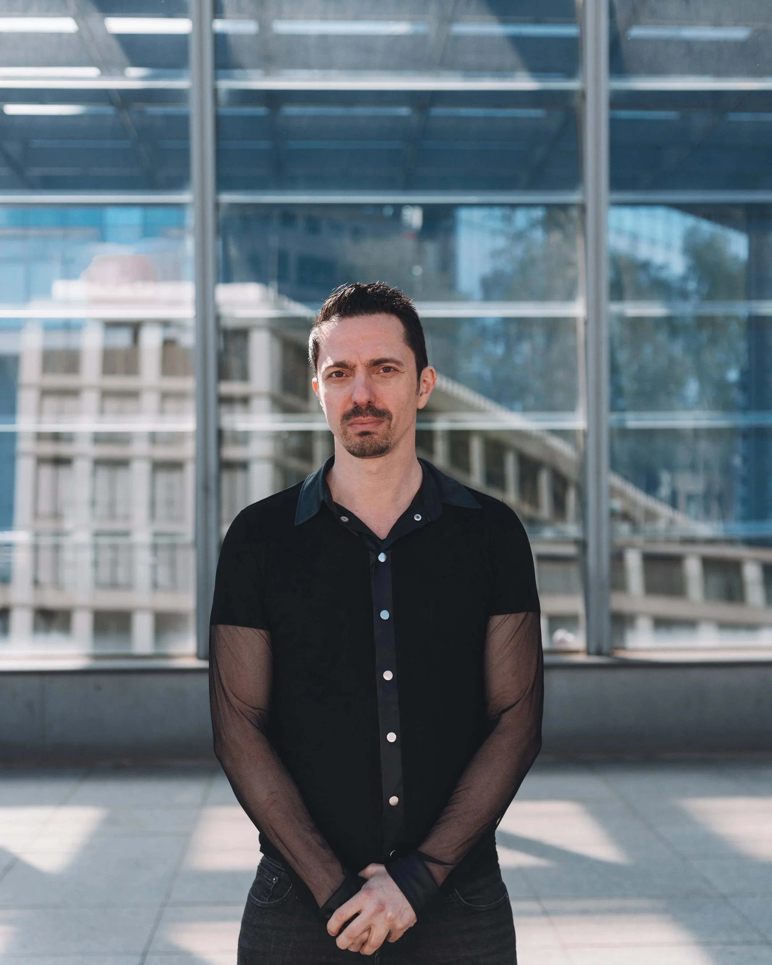 A man standing outside in front of a modern building with large glass windows, wearing a black shirt with transparent mesh sleeves.