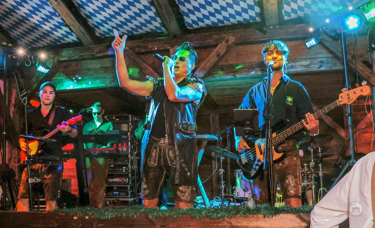 A band performing on stage with four members, including a singer in the center wearing sunglasses and a leather vest, a guitarist on the left, a keyboard player in the back, and a bass player on the right, all in a rustic setting with wooden walls an