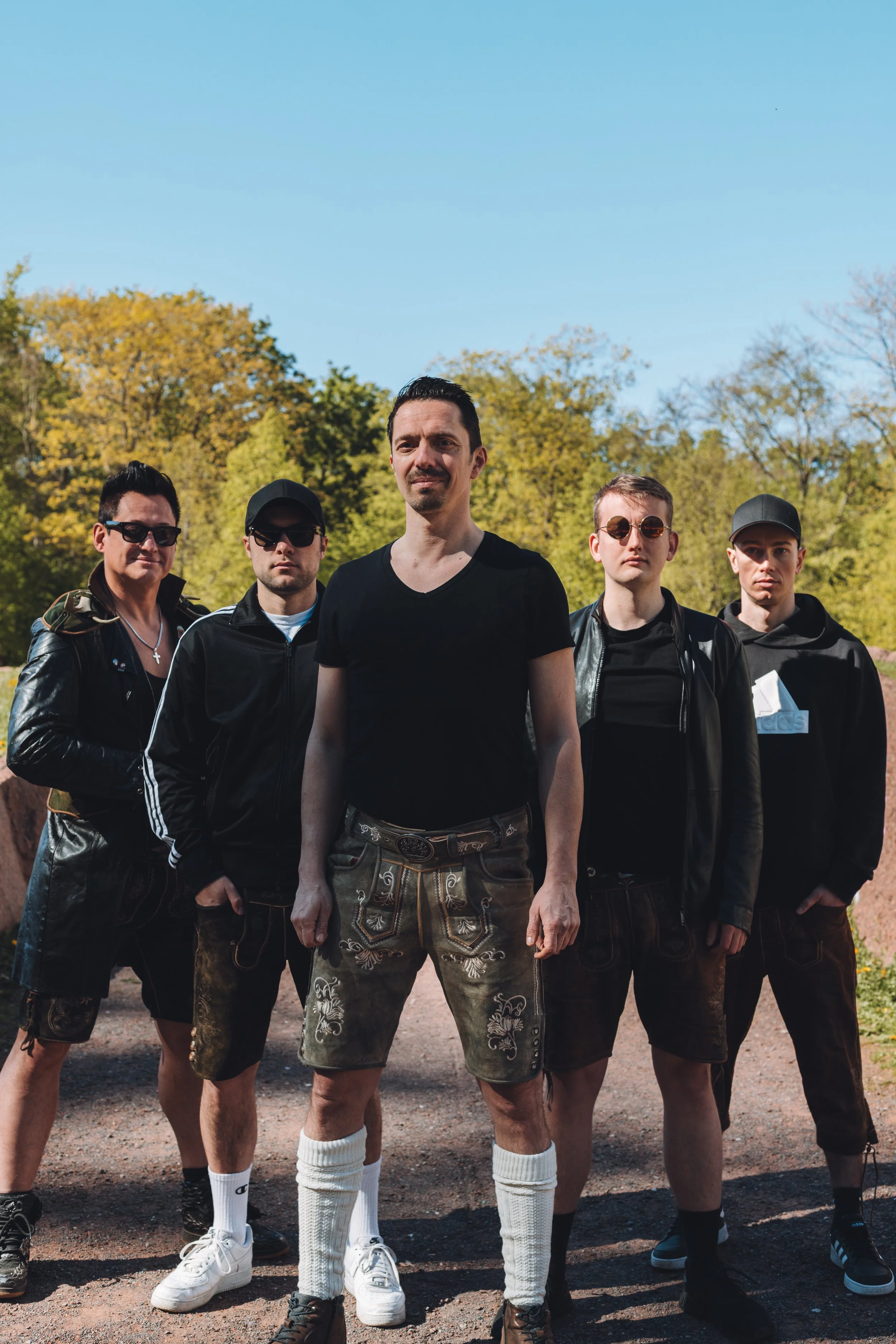 A group of five men standing outdoors in a park with trees and blue sky, dressed in casual and traditional clothing, with serious expressions.