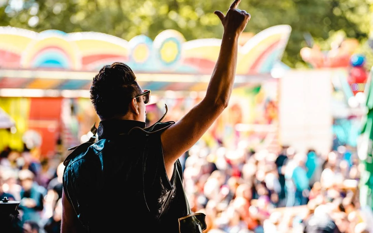 Person with short hair wearing sunglasses and a leather vest at a fair or carnival.