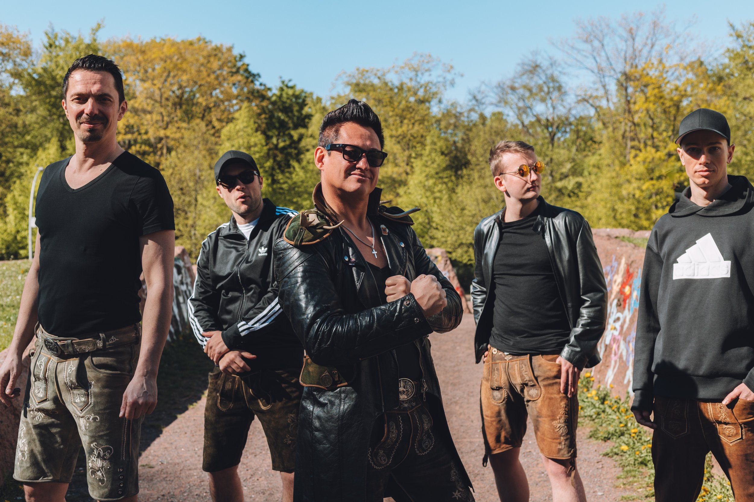 Group of five men standing outdoors on a sunny day, dressed in casual and leather clothing, with trees and a blue sky in the background.