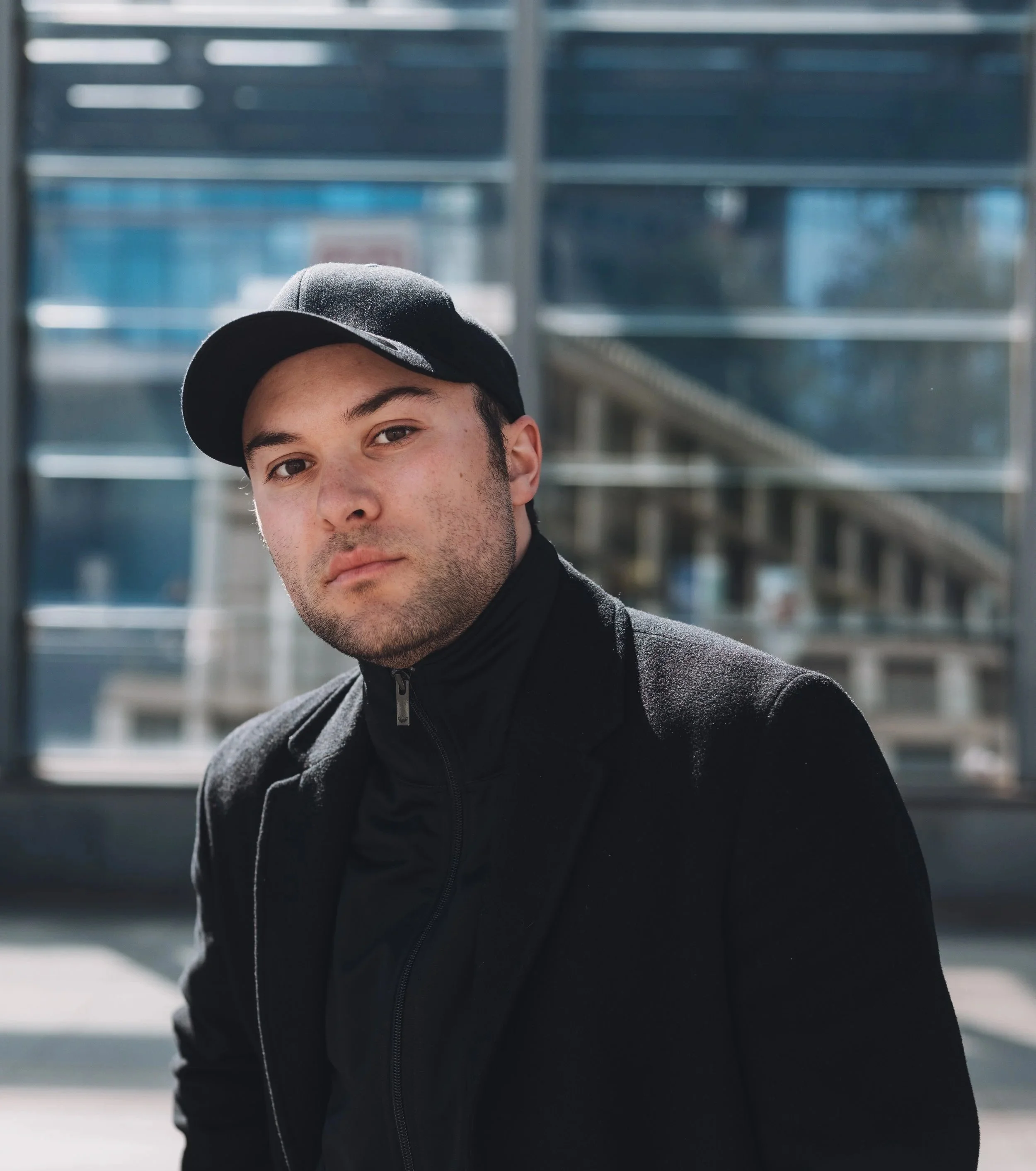 Close-up of a young man wearing a black baseball cap and black jacket outdoors with blurred modern glass building backdrop.