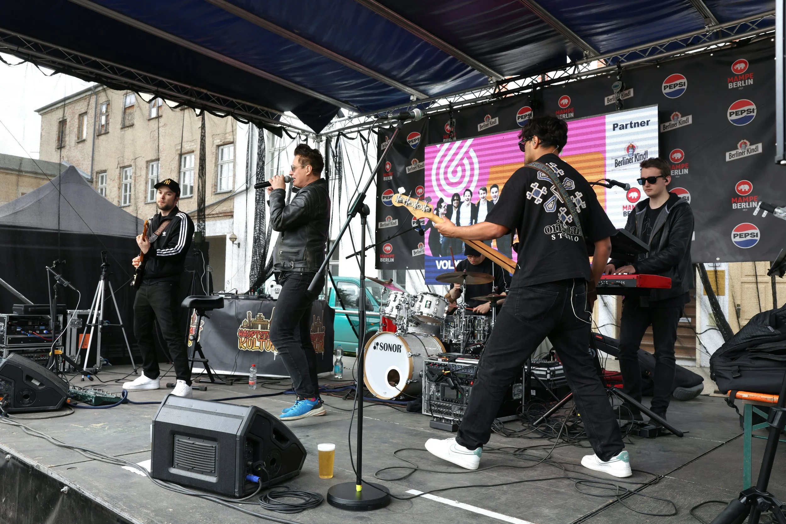 A live outdoor band performance on a stage with four musicians playing instruments and singing. The backdrop features logos of Pepsi, Berliner Qualität, and Mampfe Berlin, with a digital screen displaying a group photo and text. The stage has sound e
