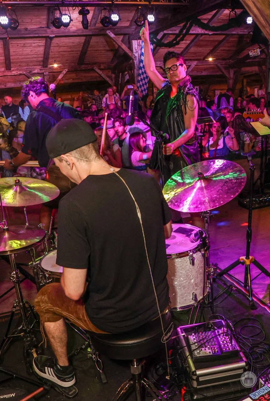 A band performs on stage in a rustic wooden venue with an audience watching. One musician is on drums, another is singing and holding a flag, and crowd members are dressed in casual and traditional attire.