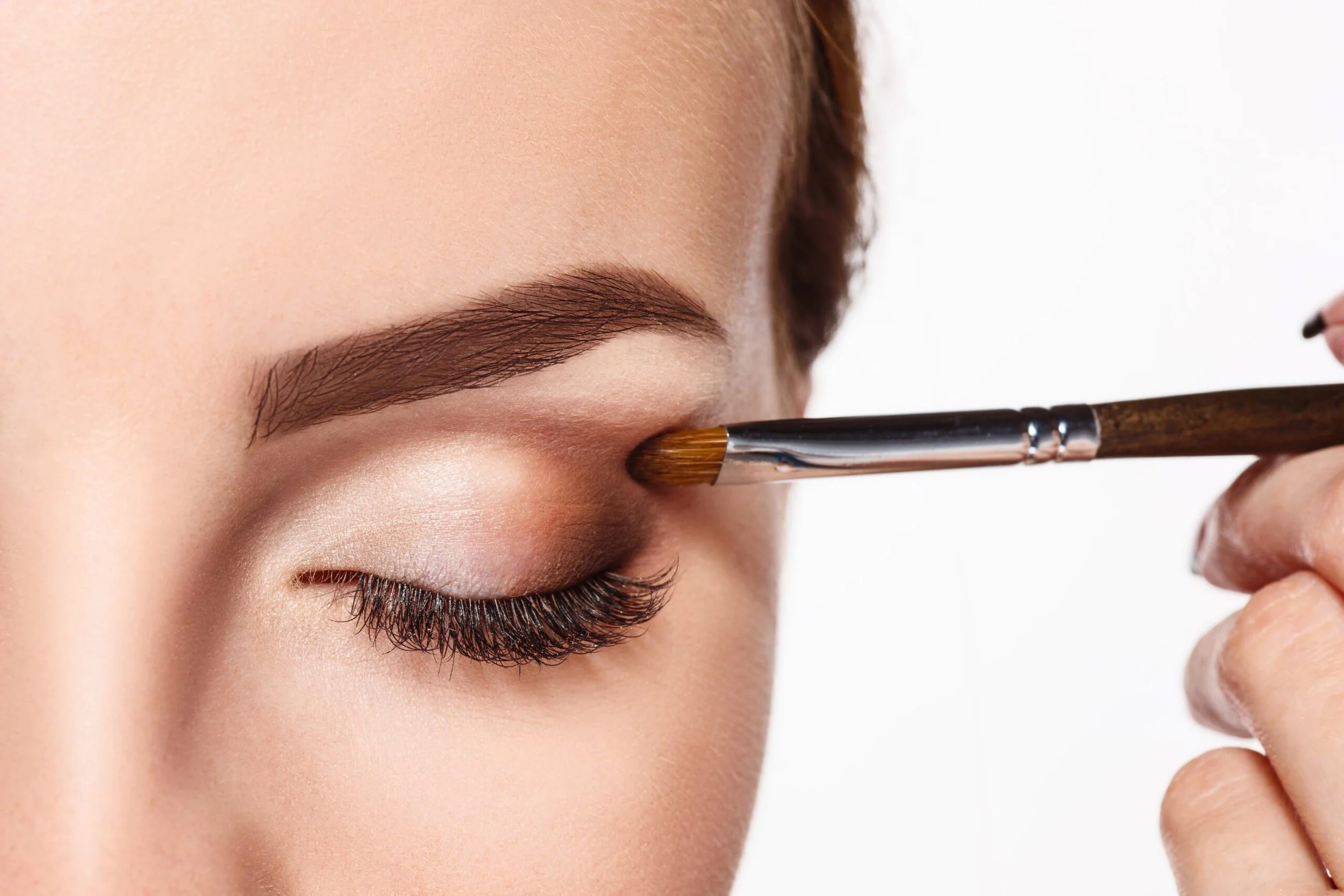 Close-up van een vrouw met gesloten ogen terwijl haar wenkbrauw wordt gevleekt met een penseel, huid en make-up worden aangebracht.