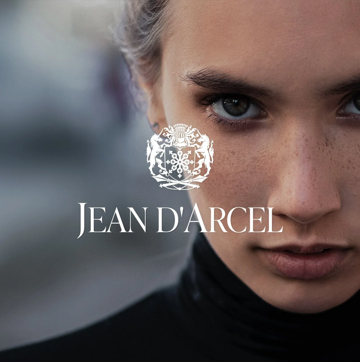 Close-up van een meisje met blauwe ogen en lichte huid, met de tekst 'Jean D'Arcel' en een grafische versie van een wapenschild.