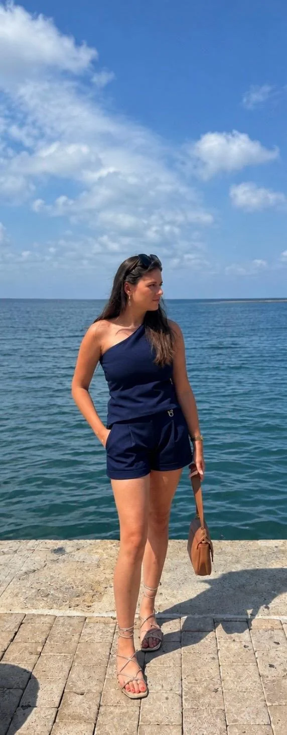 Vrouw in een navy blauw outfit staat aan de rand van het water onder een blauwe lucht met witte wolken.