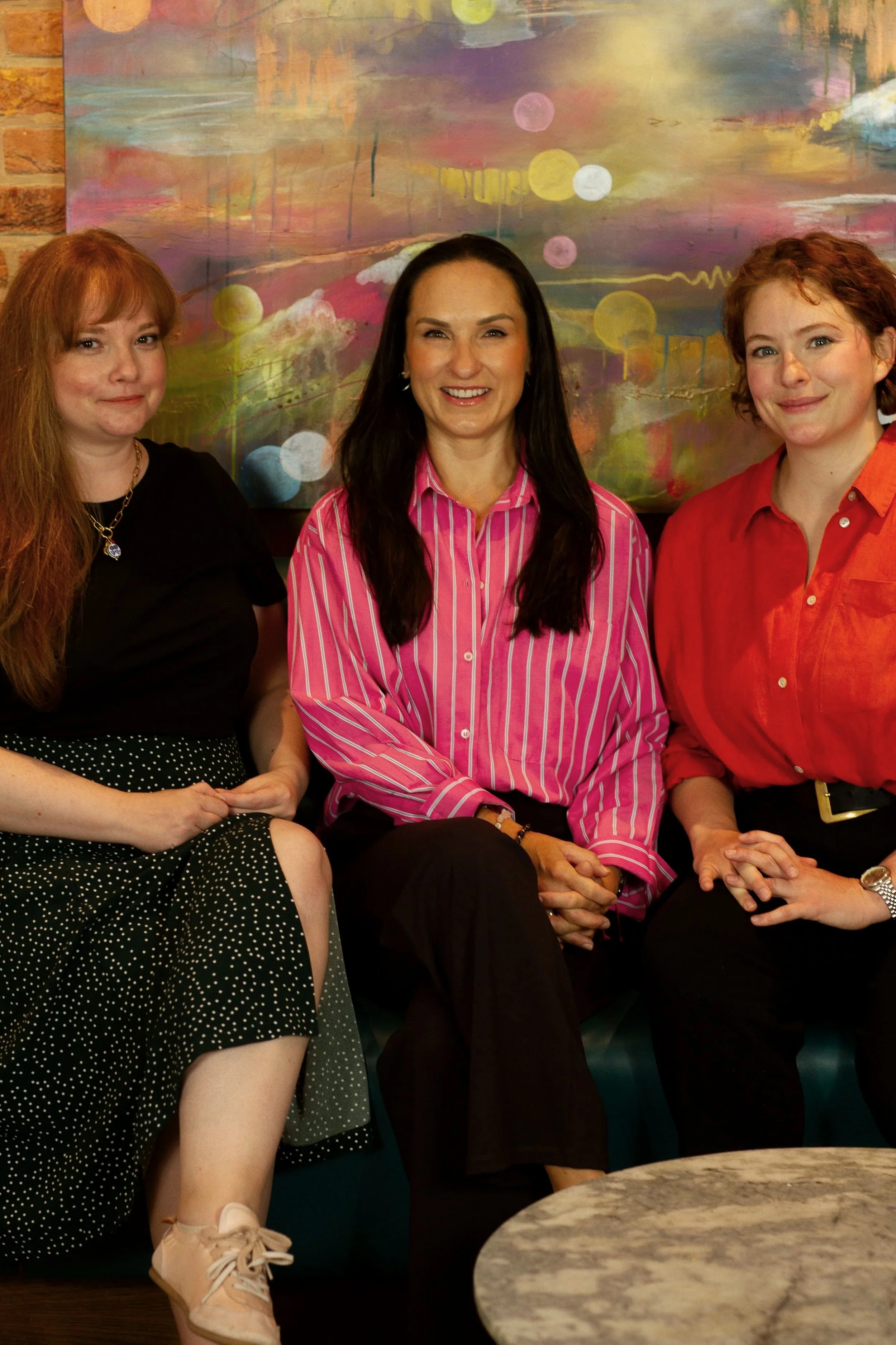 Three women sitting together in front of a colorful abstract painting. One woman has long red hair and is wearing a black top and polka dot skirt. The woman in the middle has dark hair, wearing a pink striped shirt. The third woman has curly red hair, wearing a red shirt. They are smiling and sitting in a cozy indoor setting.
