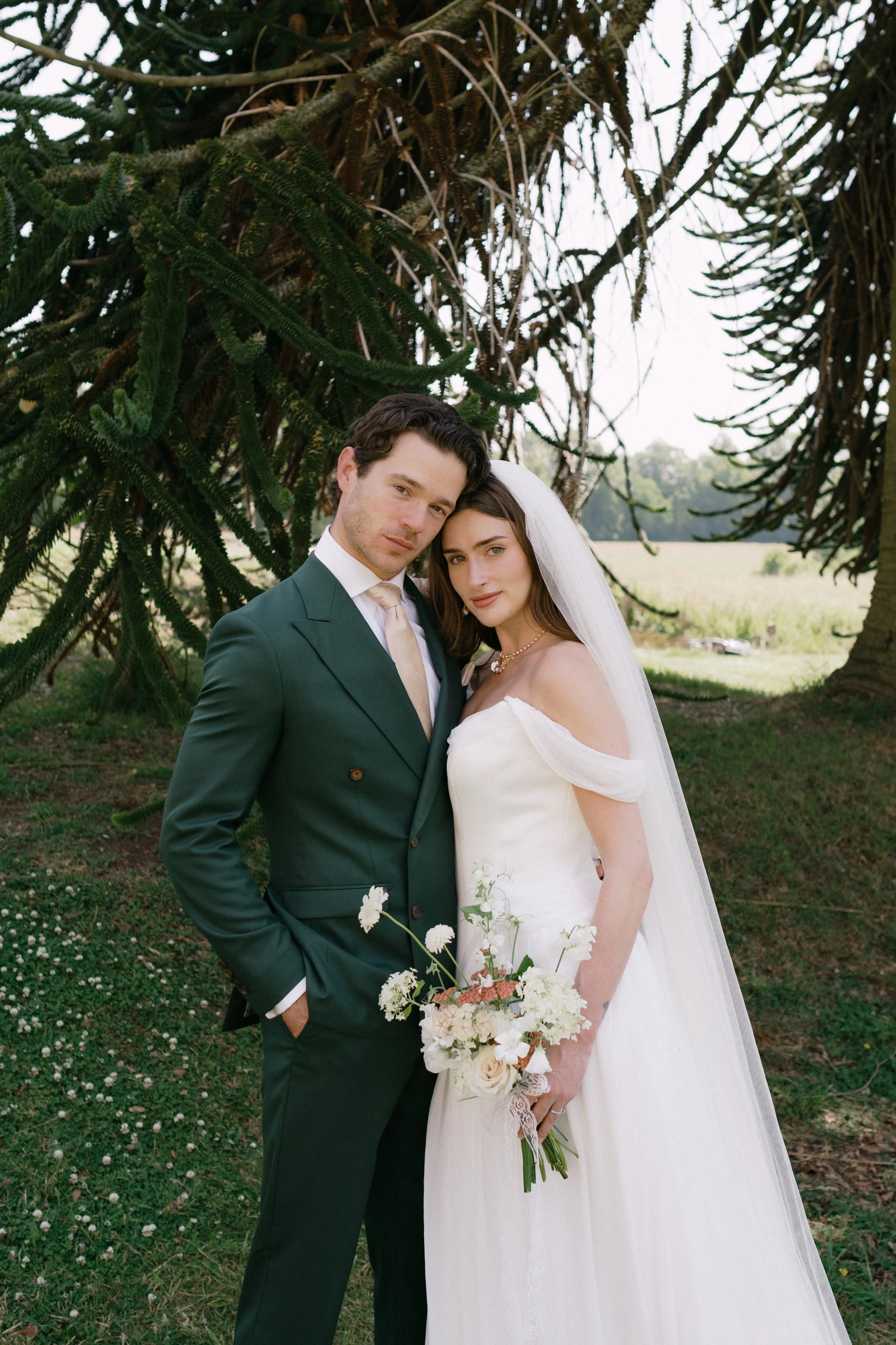 A bride and groom standing close together outdoors under a large tree. The groom is wearing a dark green suit, and the bride is in a white wedding dress holding a bouquet of white and peach flowers. The bride has a veil and a pearl necklace, and both are looking at the camera.