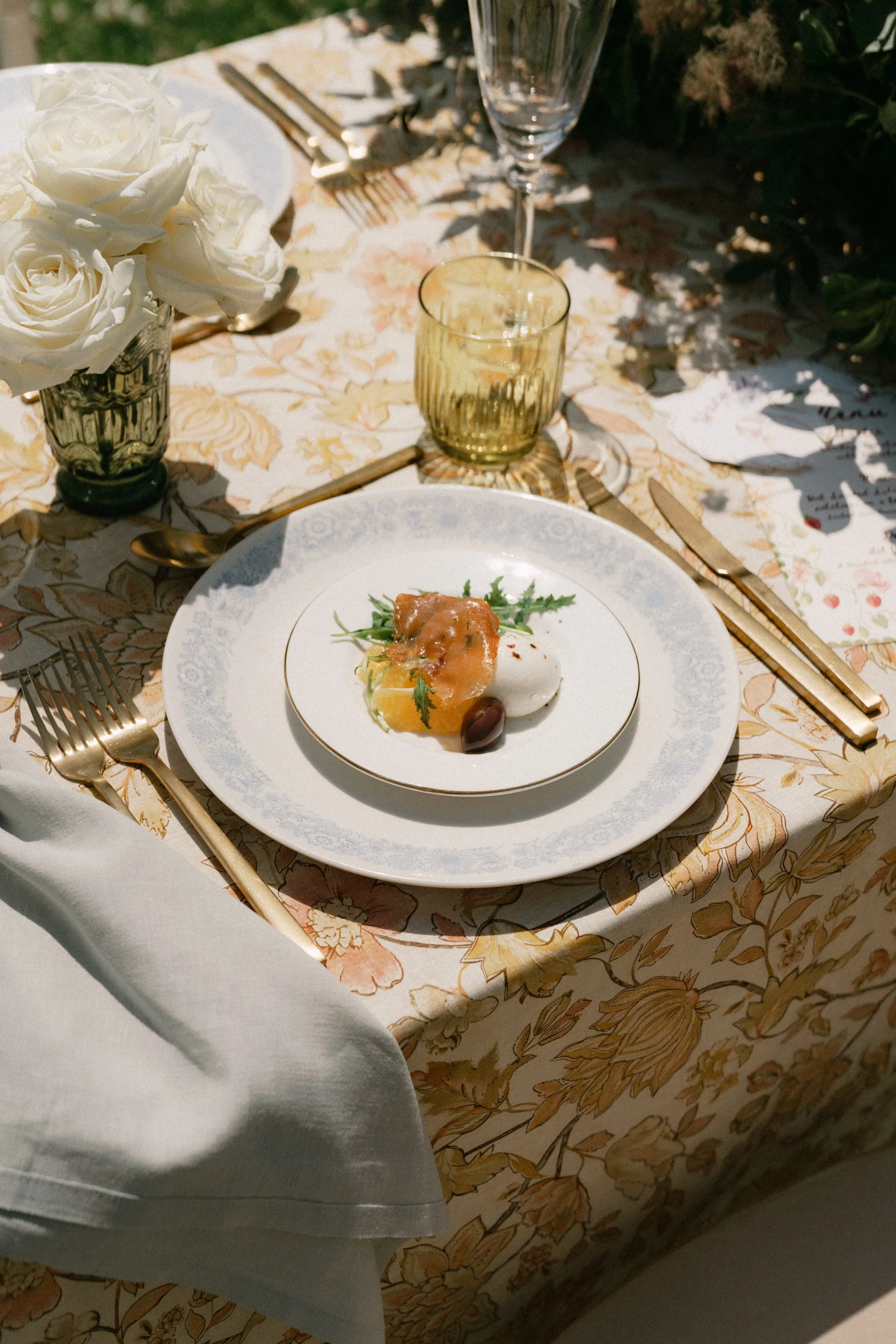 Elegant outdoor dining table set with white roses in a glass vase, a white plate with food, gold utensils, a gold-rimmed glass, a clear wine glass, and decorative tablecloth with a floral pattern.