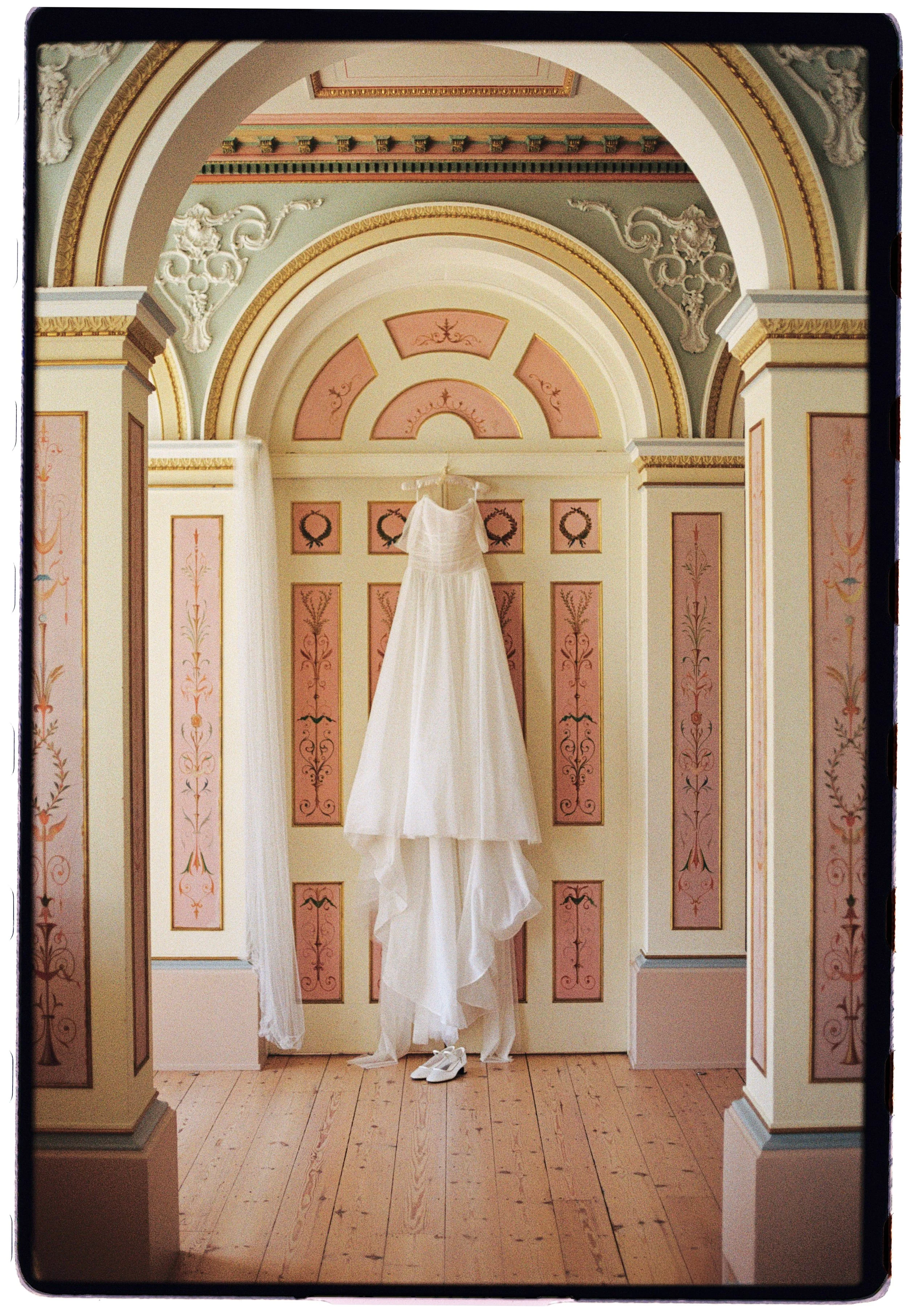 A white wedding dress hanging on a hook in a decorated room with pastel pink and cream walls and intricate moldings, with a pair of white shoes on the floor below.