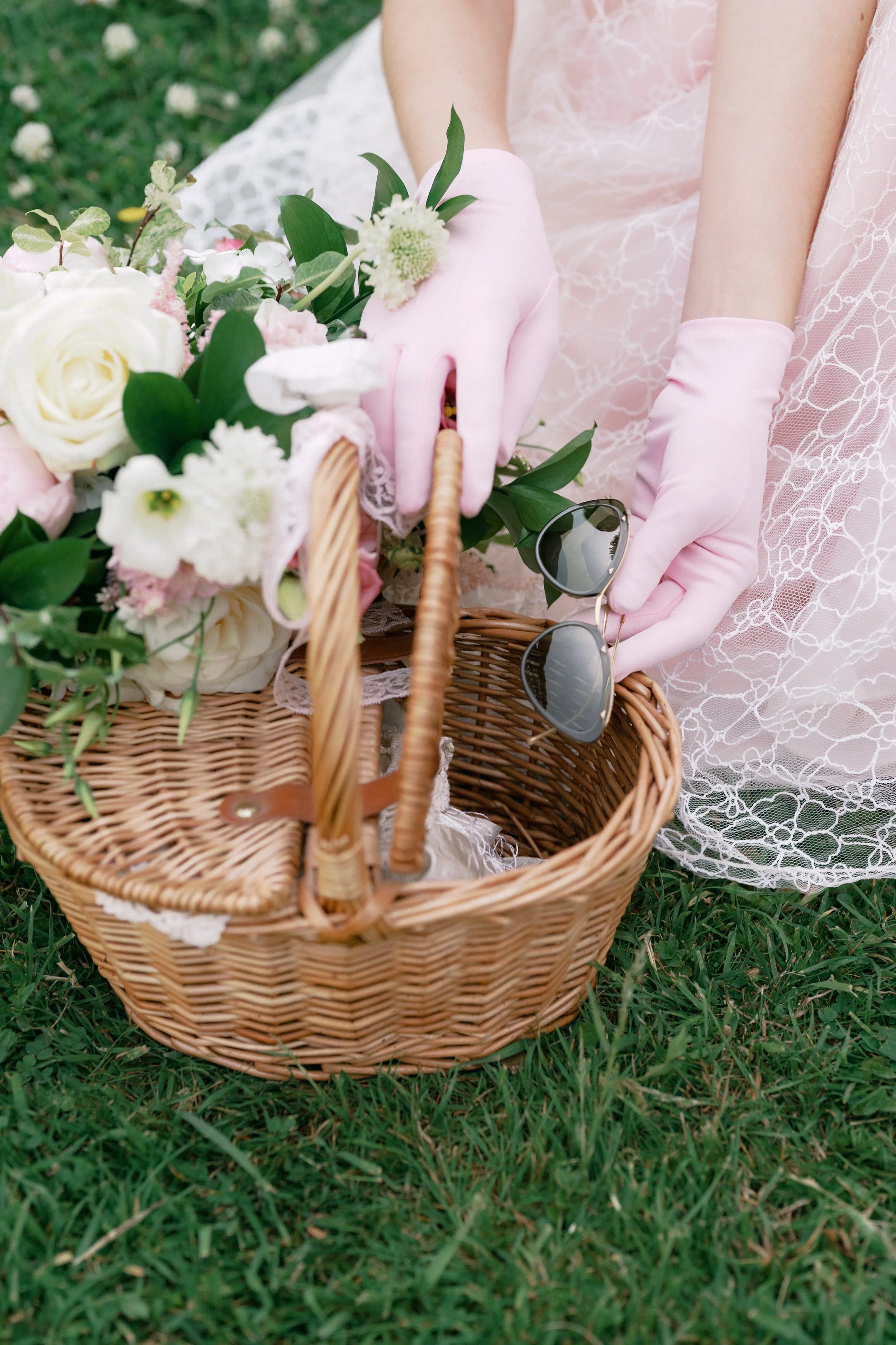 A woman wearing pink lace gloves and a pink lace dress is placing her sunglasses into a wicker picnic basket filled with white and pink flowers. She is sitting on green grass outdoors.