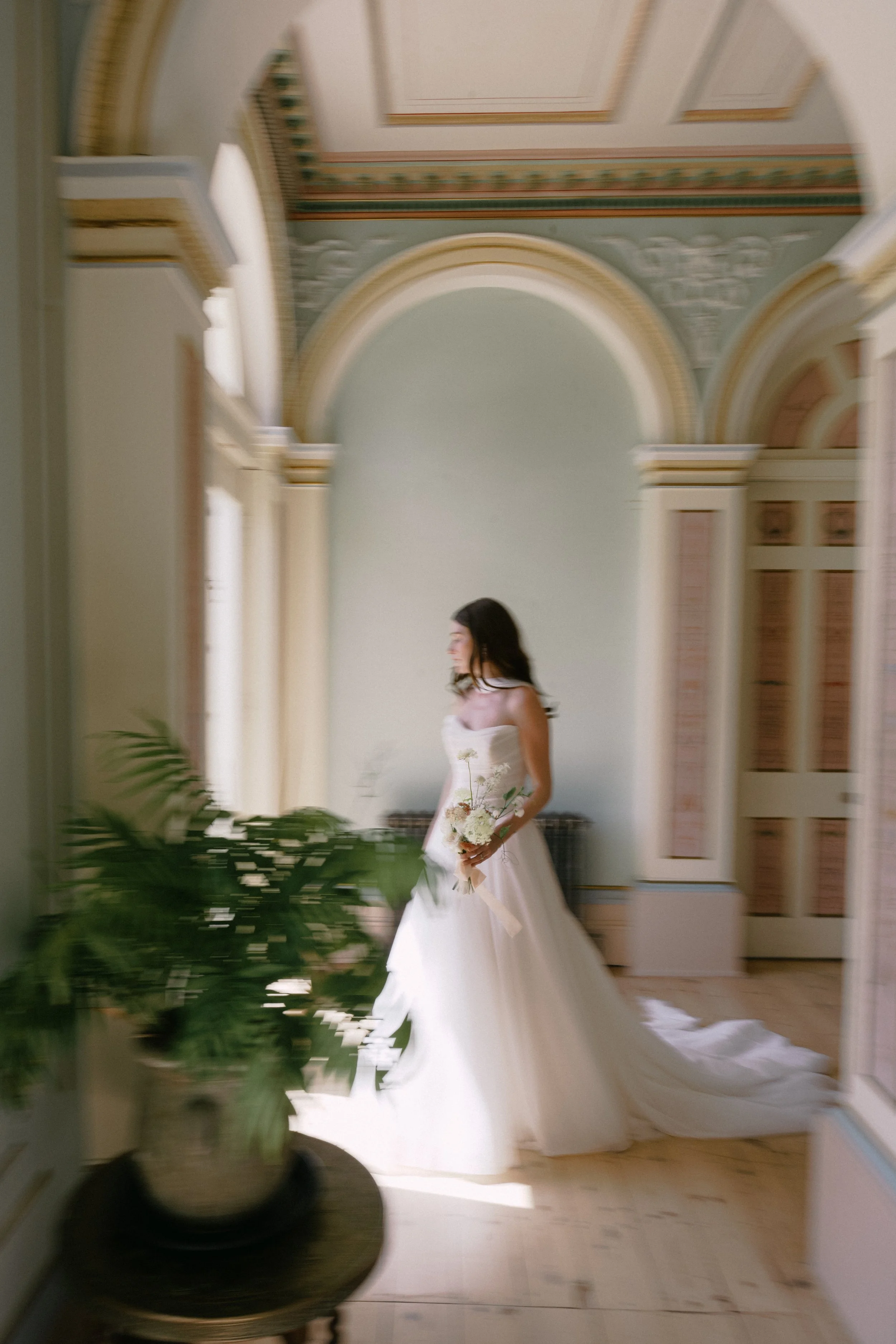 A woman in a wedding dress holding a bouquet standing in an ornate interior room with archways and detailed trim.