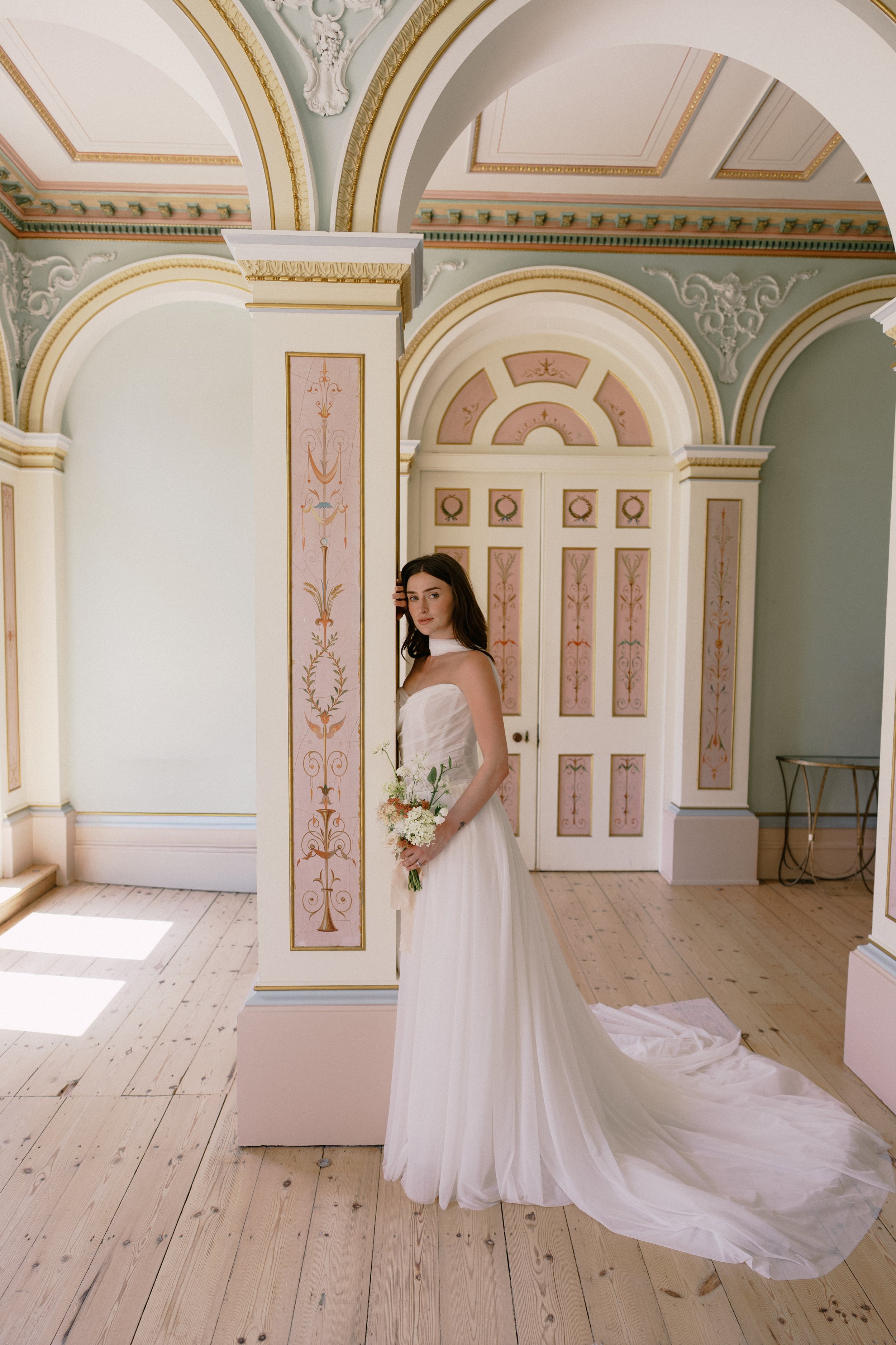 A woman in a white strapless wedding gown holding a bouquet of white flowers, standing in an ornate room with pastel pink and green decor, gold accents, and arched ceilings.