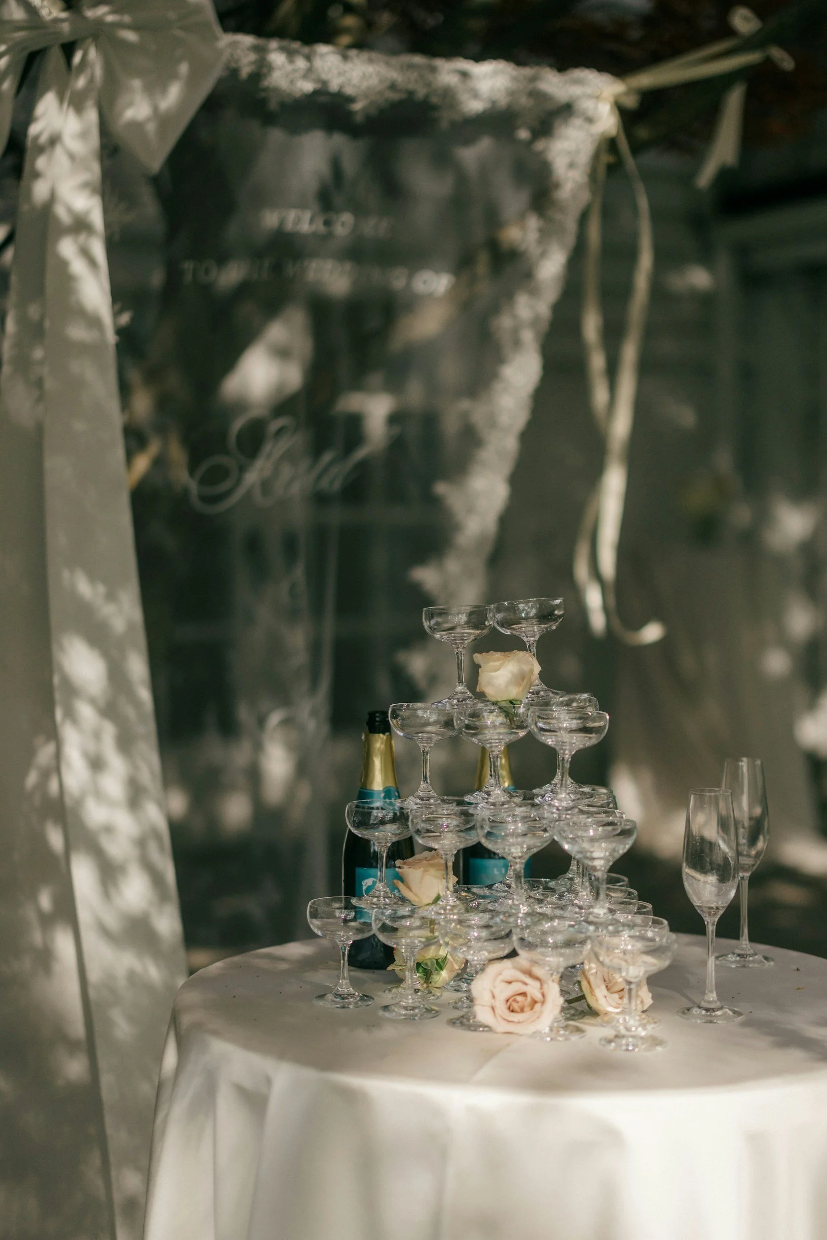 A champagne tower with glasses stacked in a pyramid shape on a round table covered with a white cloth, decorated with roses, with bottles of champagne nearby. The background shows draped fabric and a large chair.