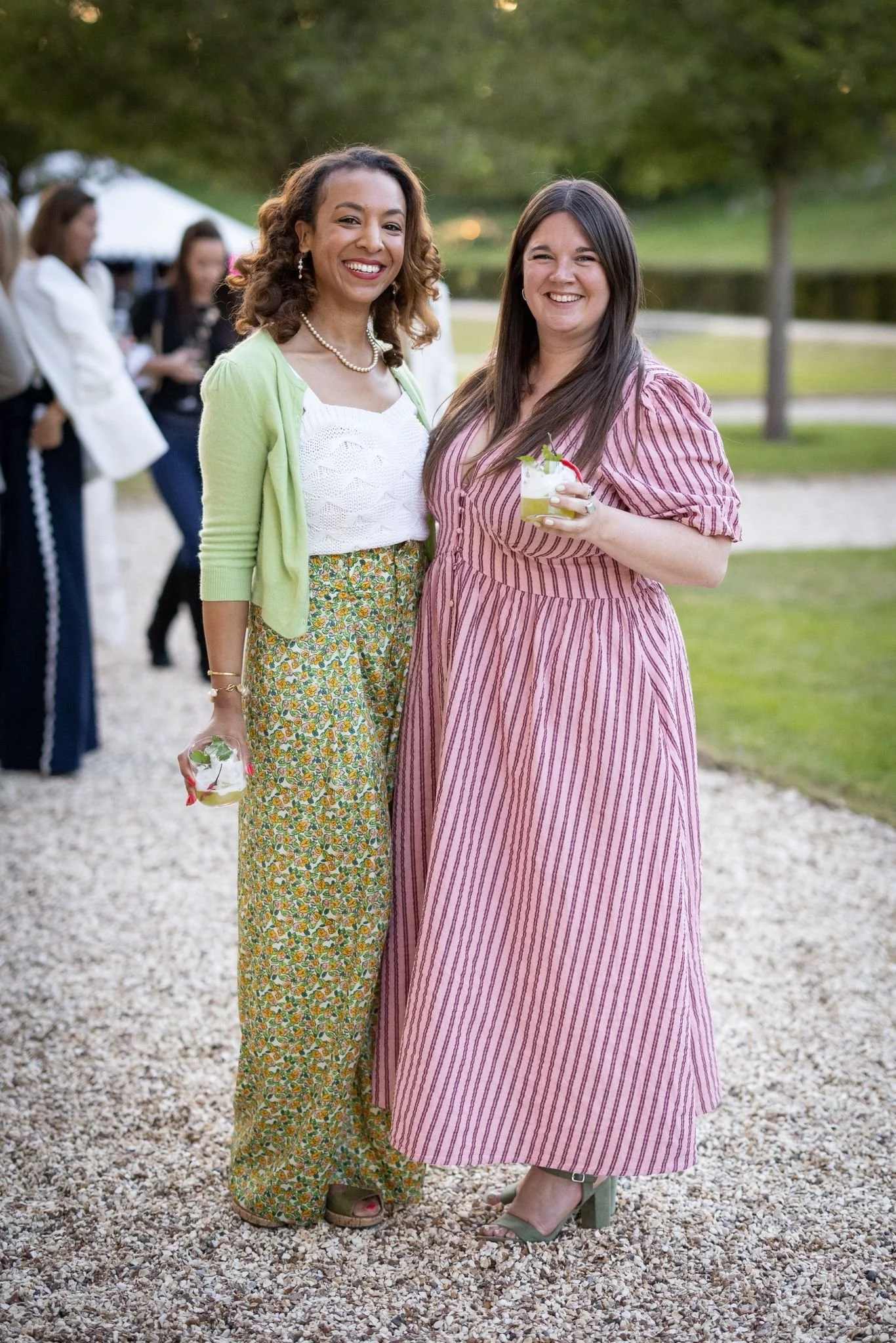 Two women smiling outdoors, holding drinks, dressed in colorful, patterned dresses, with blurred people and greenery in the background.