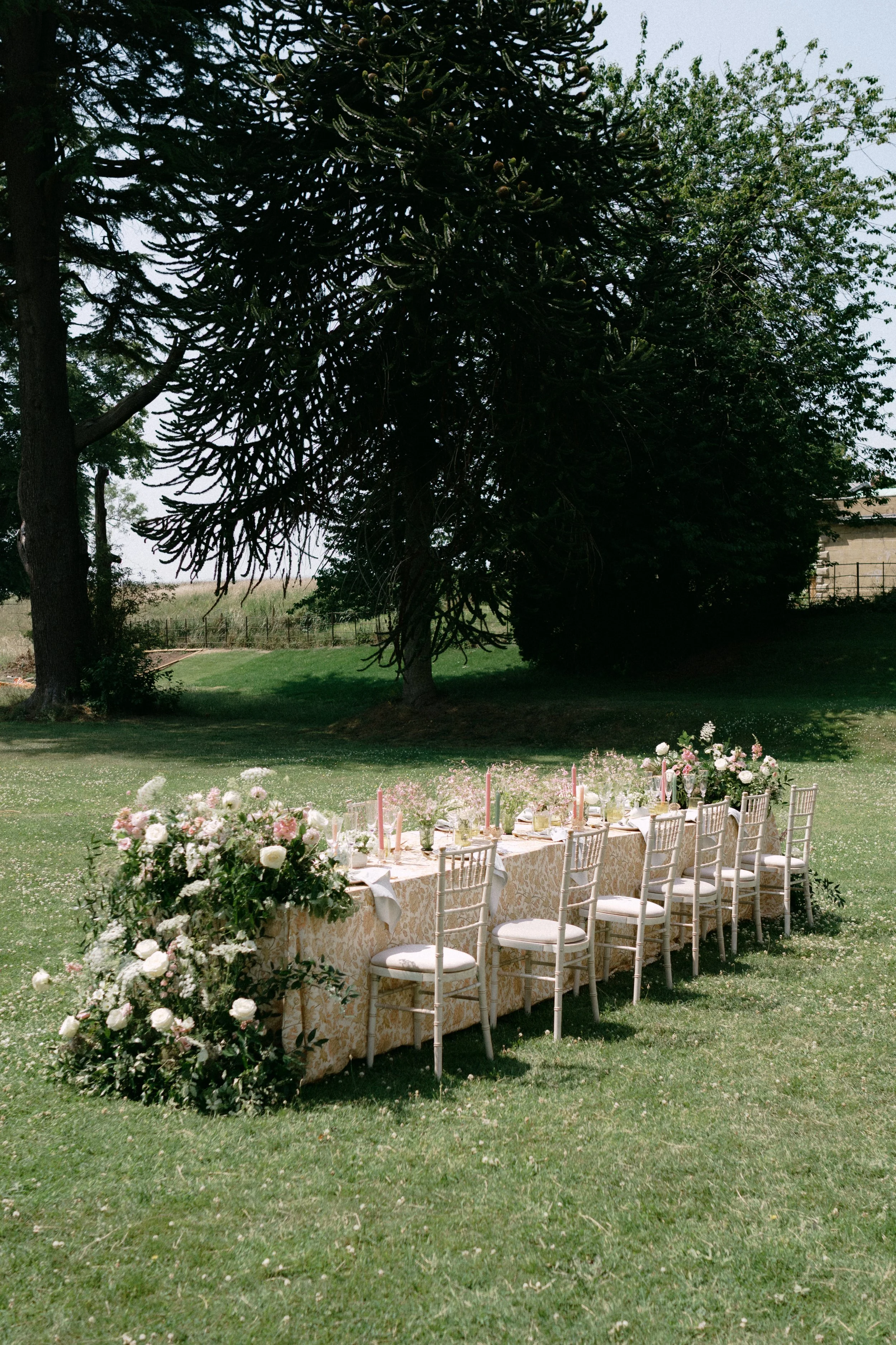 Outdoor wedding reception setup with a long decorated table surrounded by chairs, floral arrangements, and candles, set on a grassy lawn under trees.
