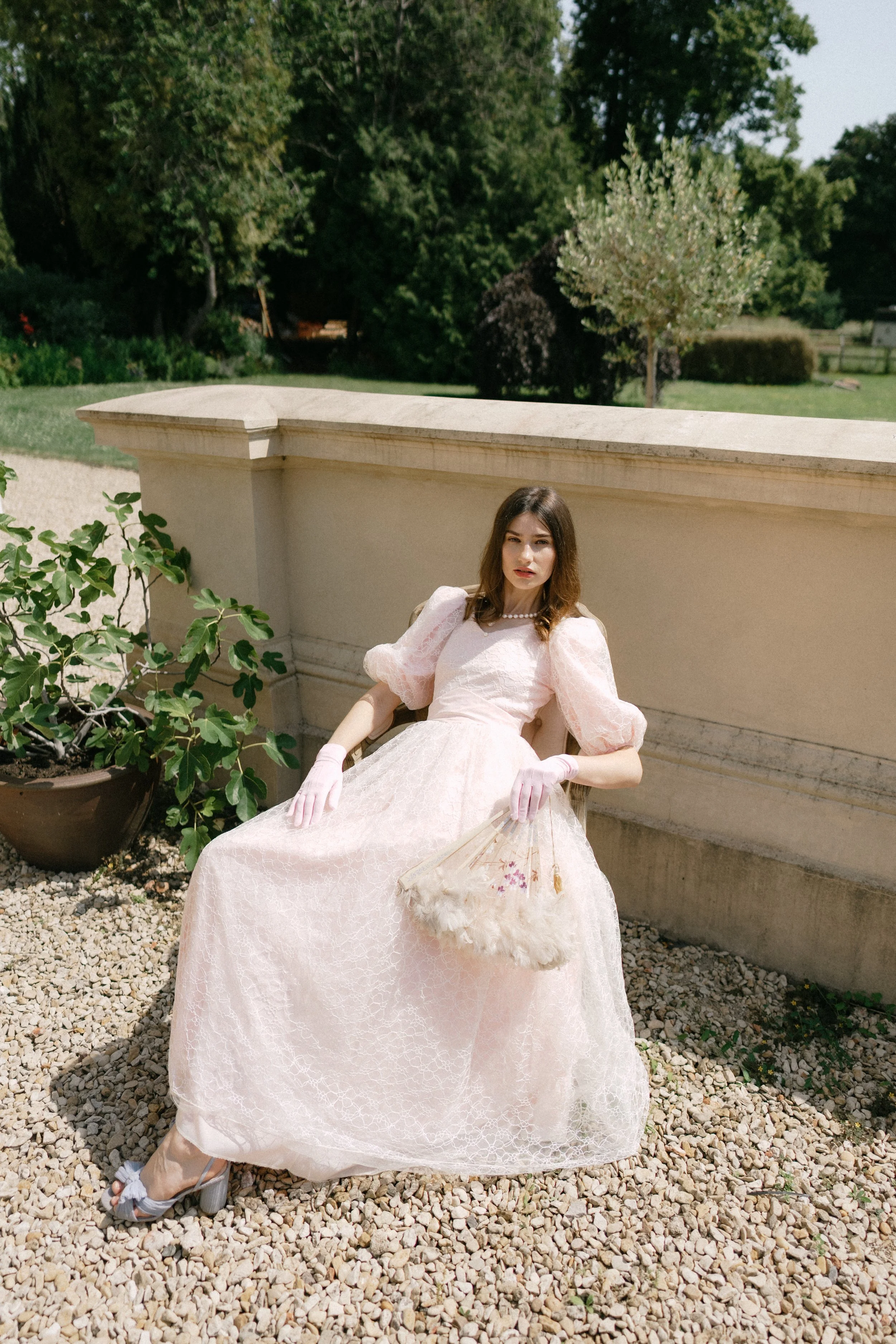 A young woman with long brown hair sitting outdoors on a gravel surface, wearing a vintage pink lace dress with puffed sleeves, white gloves, and high heeled shoes with bows. She is holding a fluffy bag and has a serious expression. Behind her is a s