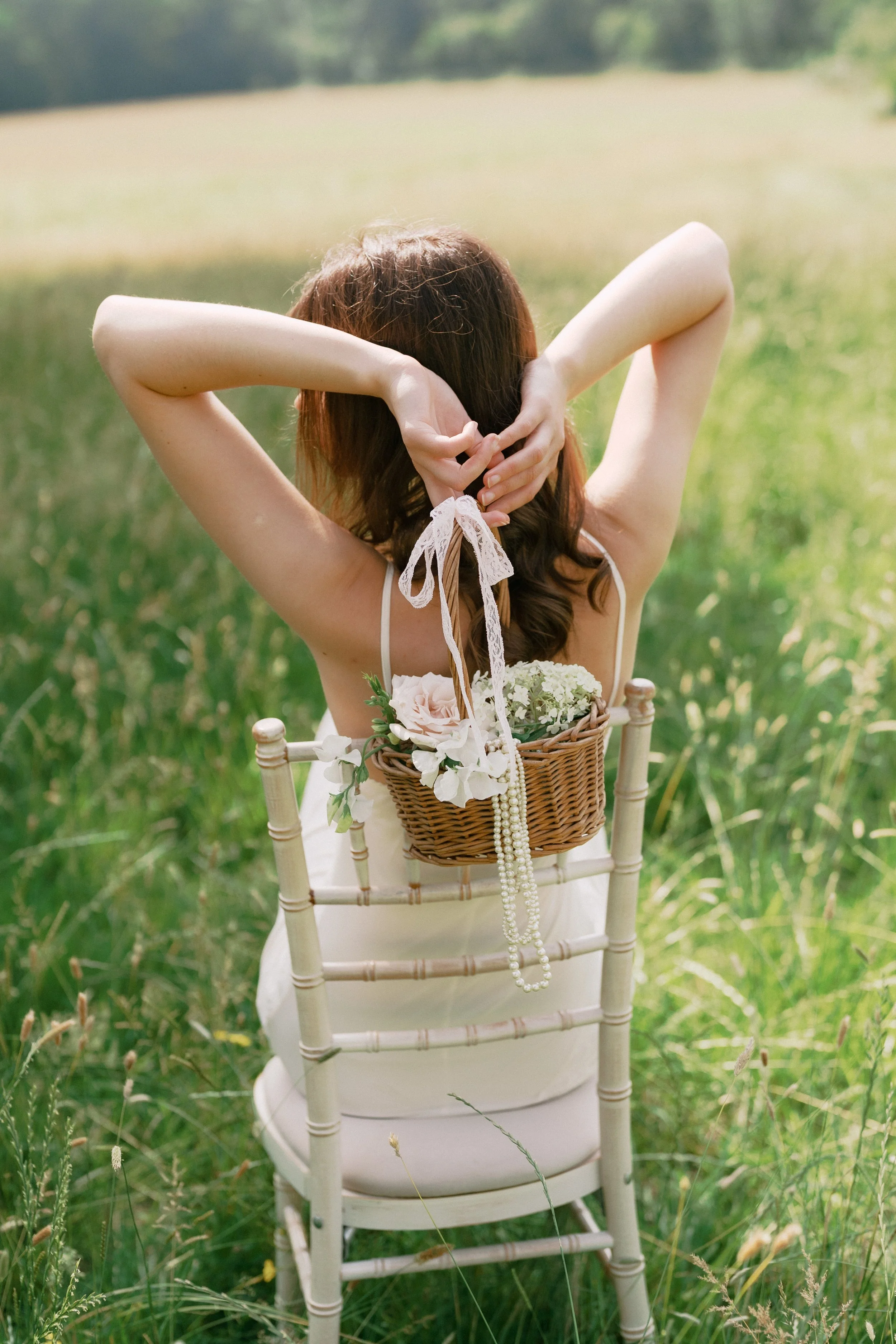 A woman with brown hair wearing a white dress sitting on a white wooden chair in a grassy field, facing away from the camera. She holds her hair up with her hands and has a basket with flowers and a pearl necklace hanging from it on her back. The background shows a blurred green landscape.