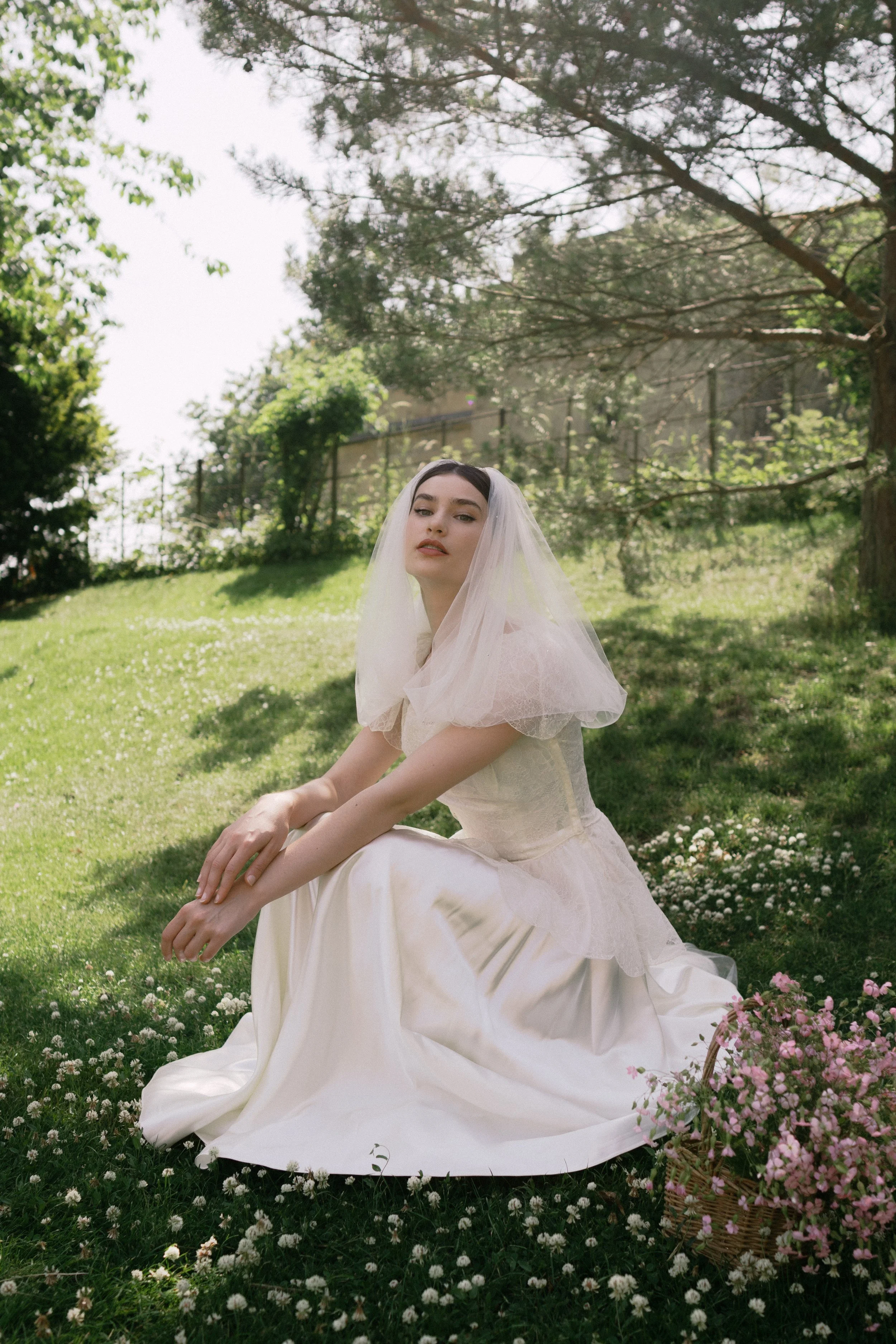 A woman wearing a wedding dress and veil sits on grassy ground in a garden, surrounded by small white and pink flowers, with trees and a fence in the background.