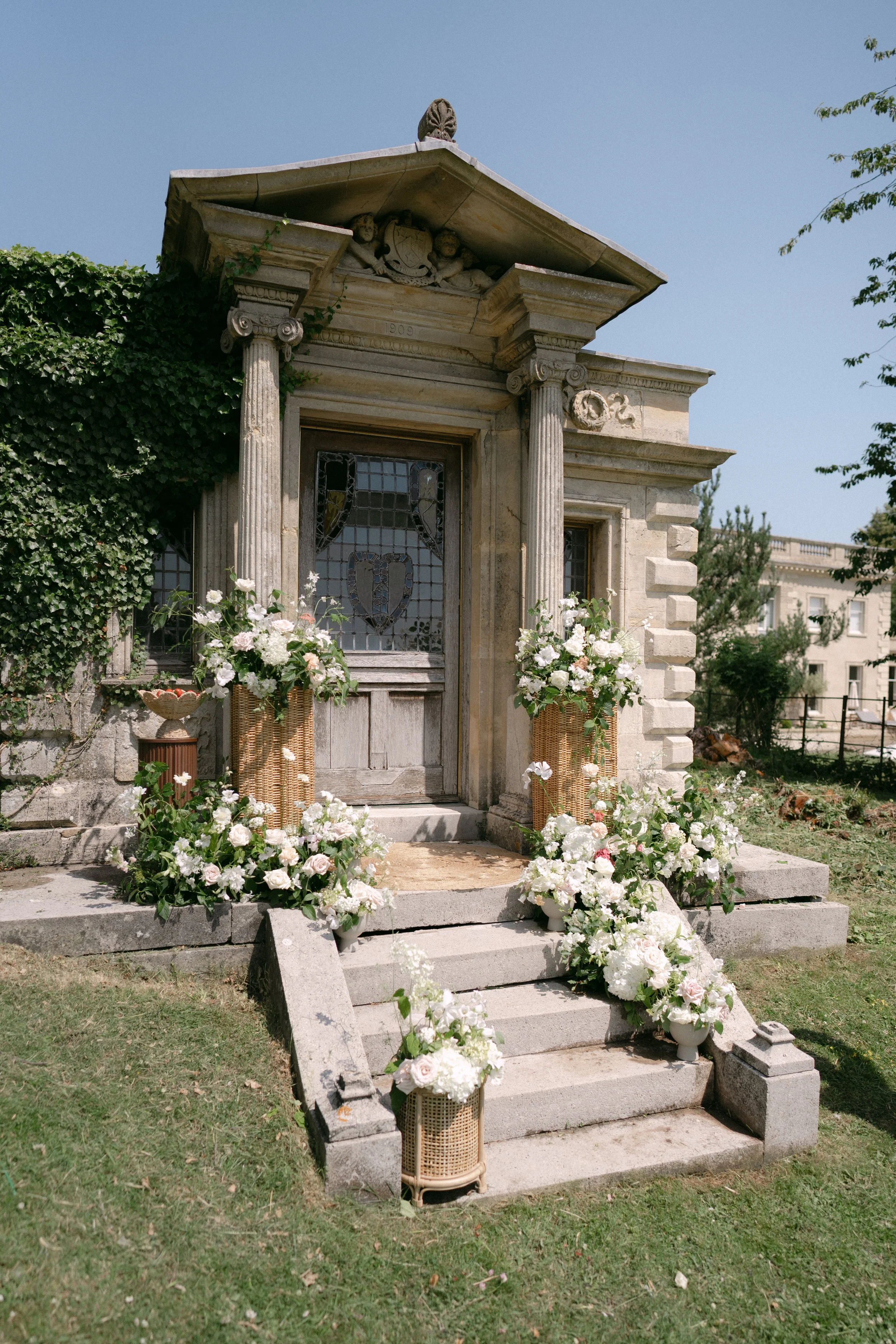 Exterior of a small stone mausoleum decorated with white and pink flowers on steps and around the entrance, with flower pots and greenery in a sunny garden setting.