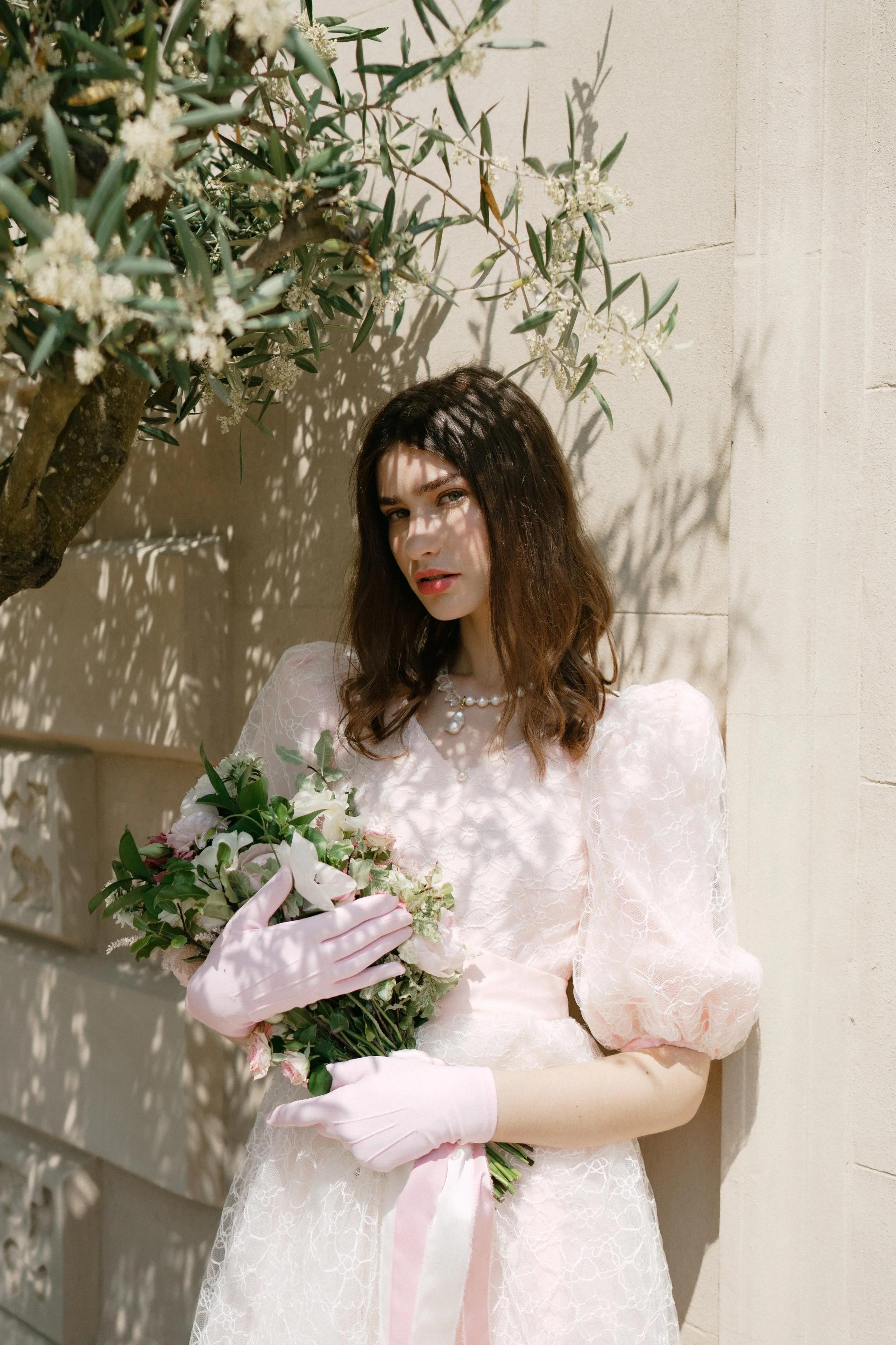 A young woman dressed in a white wedding gown holding a bouquet of flowers, standing against a light-colored wall with sunlight casting shadows from a nearby tree.