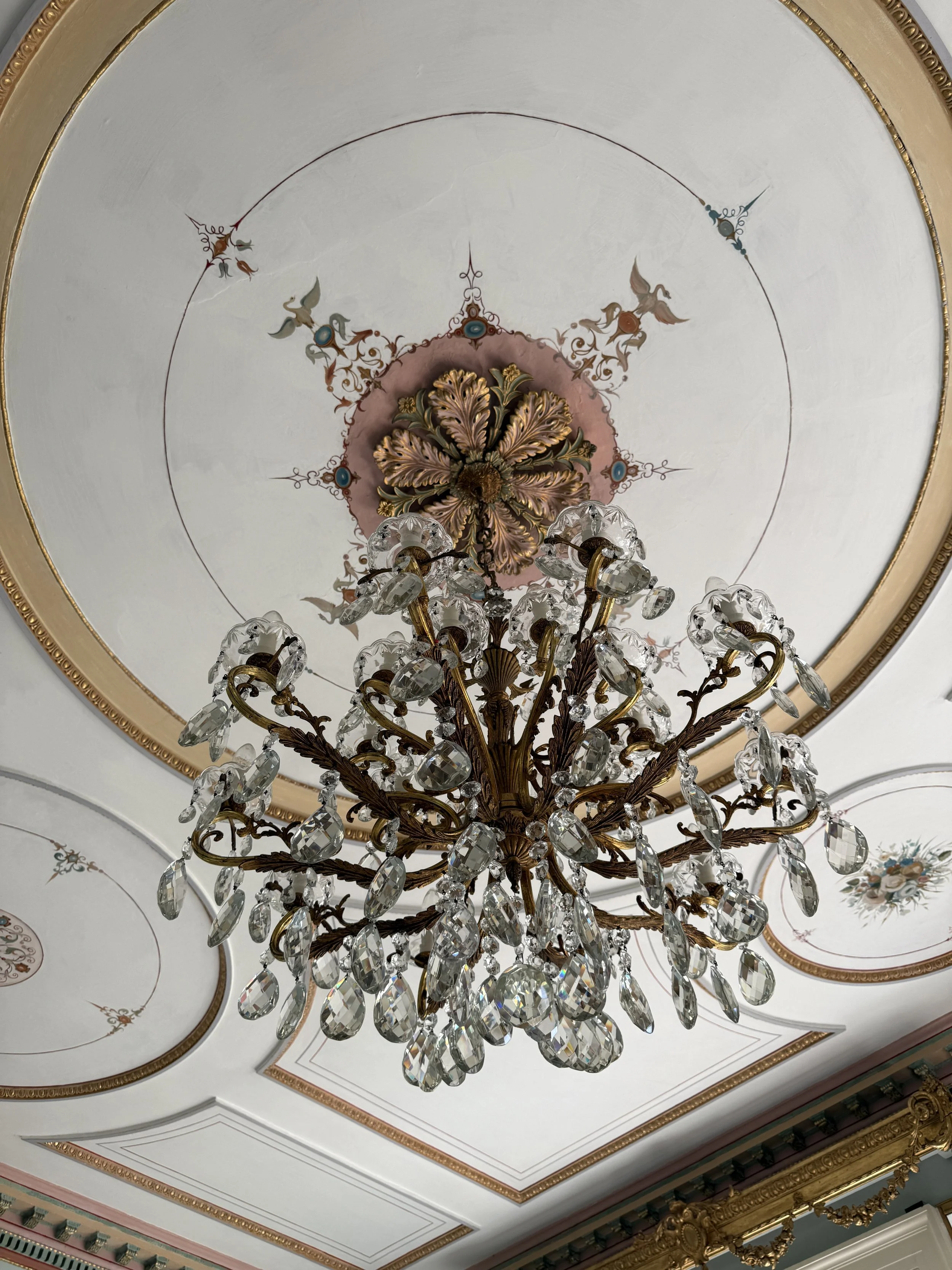 Ornate ceiling with a large chandelier made of crystal and gold, featuring intricate floral and leaf patterns, and decorative painted elements near the edges of the ceiling.