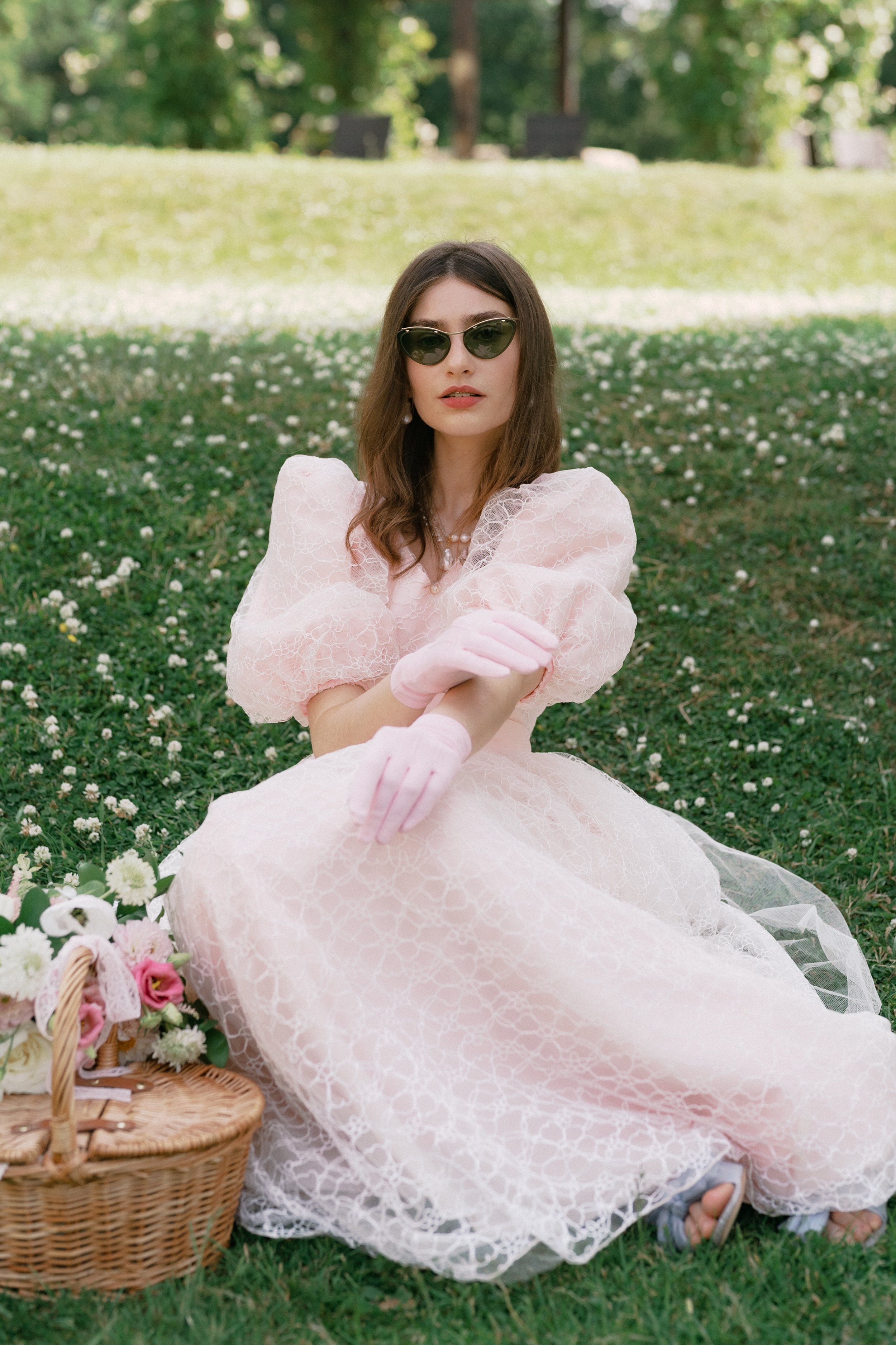 Woman wearing vintage sunglasses and a light pink lace dress sitting on grass with a picnic basket of flowers next to her, outdoors in a park-like setting with trees in the background.