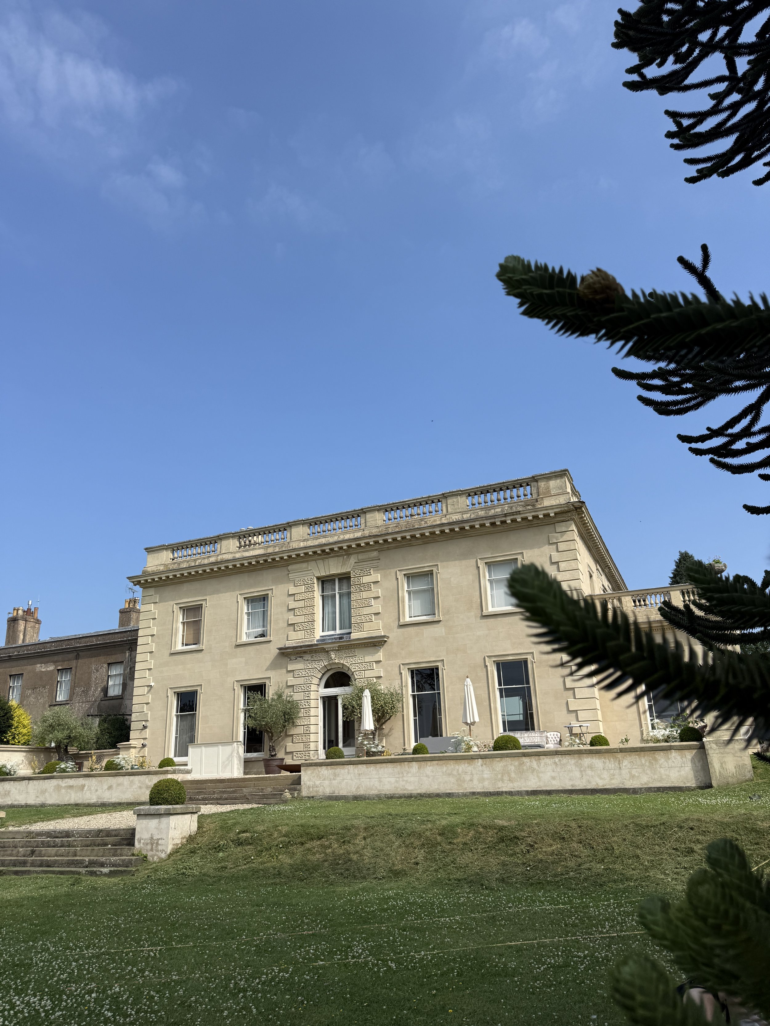 A large elegant beige building with decorative architectural details, surrounded by a well-maintained garden with small trees and shrubs, outdoor seating with umbrellas, and a stone staircase, under a clear blue sky and some leafy branches framing th