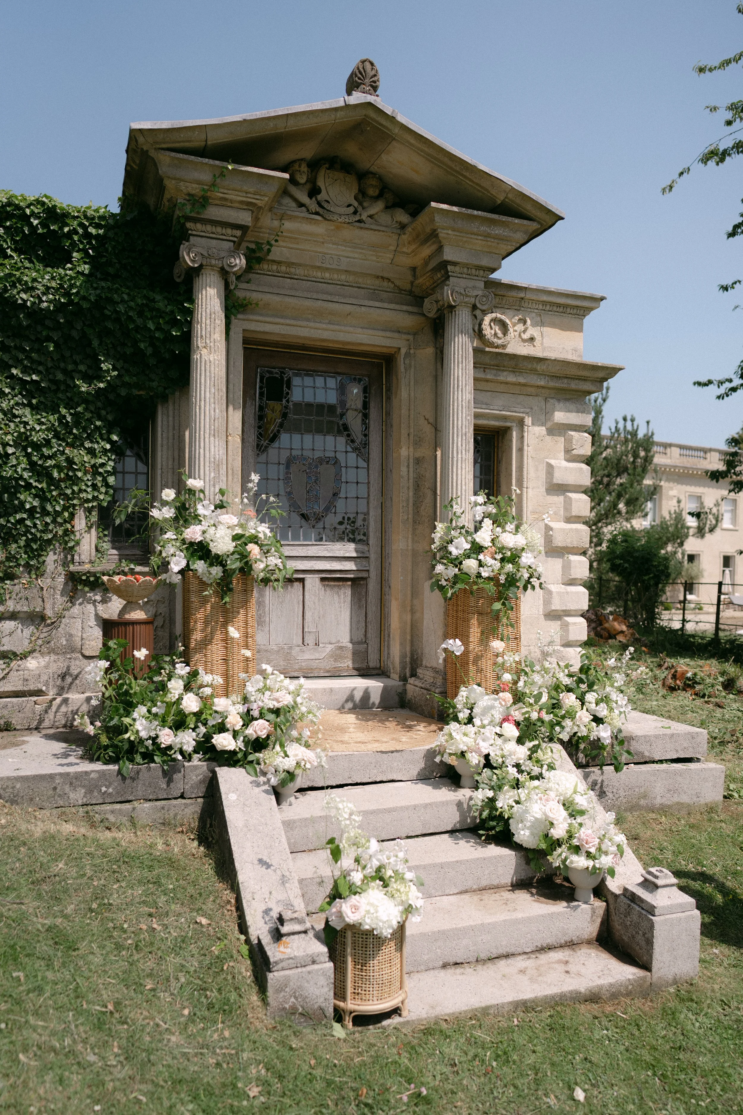 A stone mausoleum or chapel adorned with white and pink flowers arranged in baskets and vases, with steps leading up to the entrance, in a well-maintained garden.